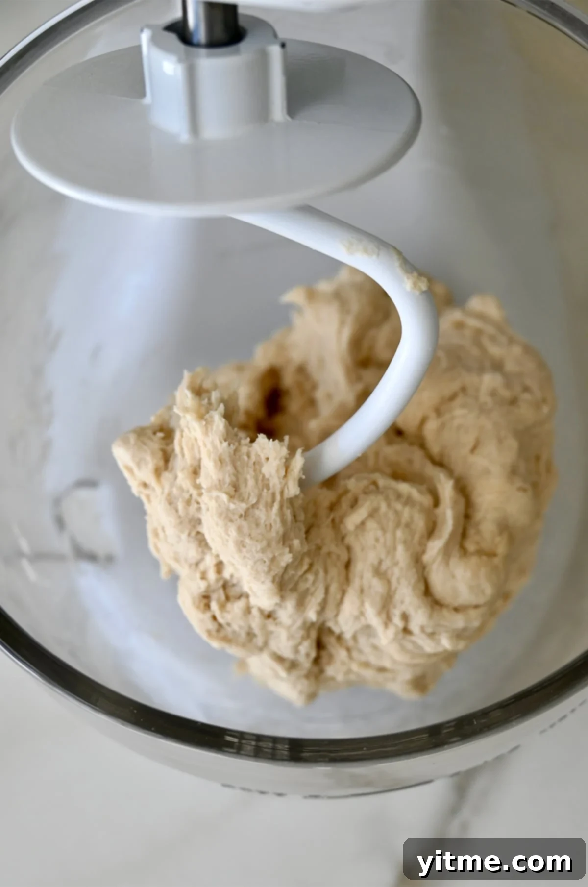Pretzel dough wrapped around the dough hook of a white stand mixer with a glass bowl.