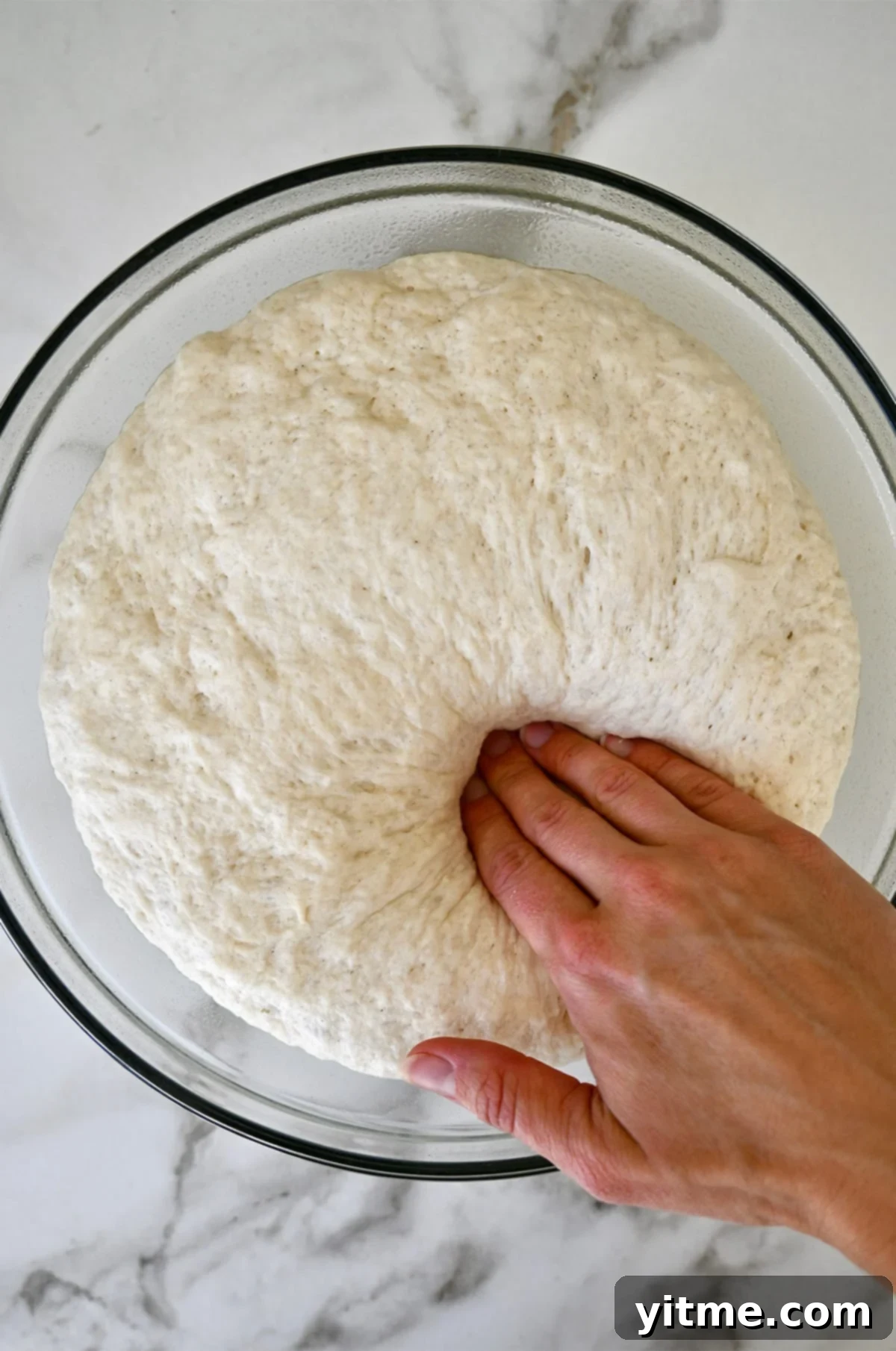 A hand presses into proofed pretzel dough in a glass bowl.