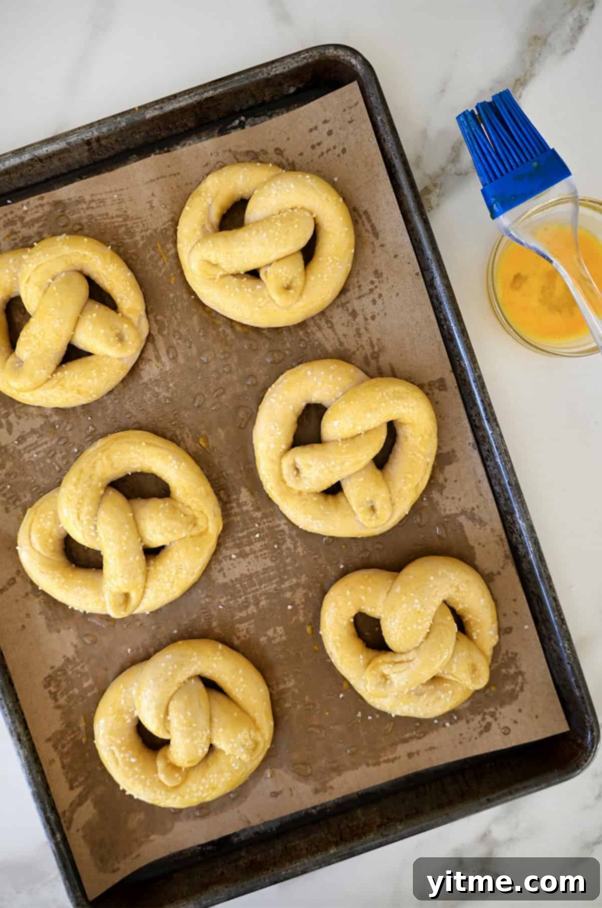 Six unbaked pretzels on a baking sheet topped with an egg wash and sprinkled with salt. A small bowl containing egg wash and a pastry brush are next to it.