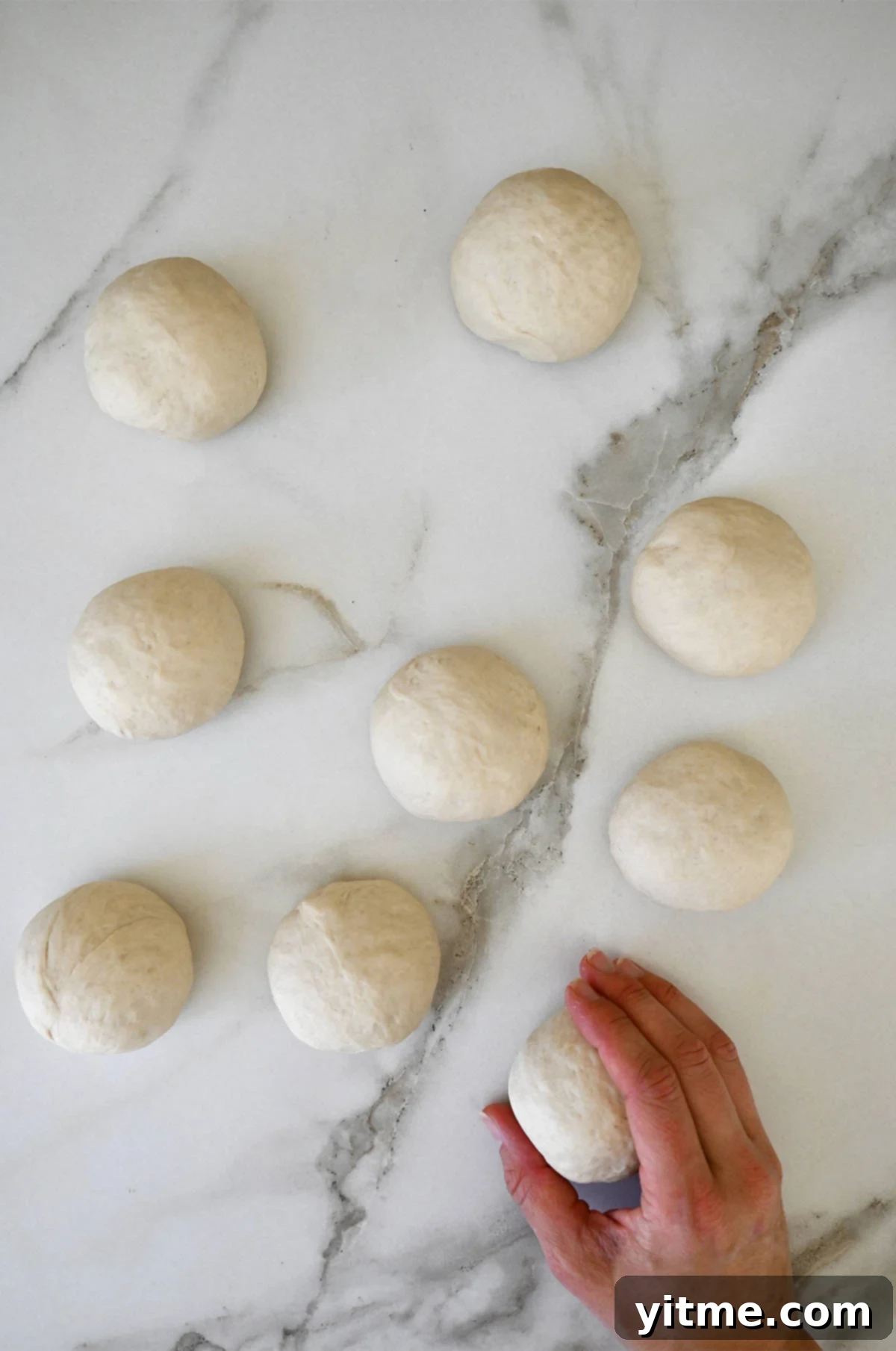 Nine dough balls on a marble surface with a hand holding onto one of them.