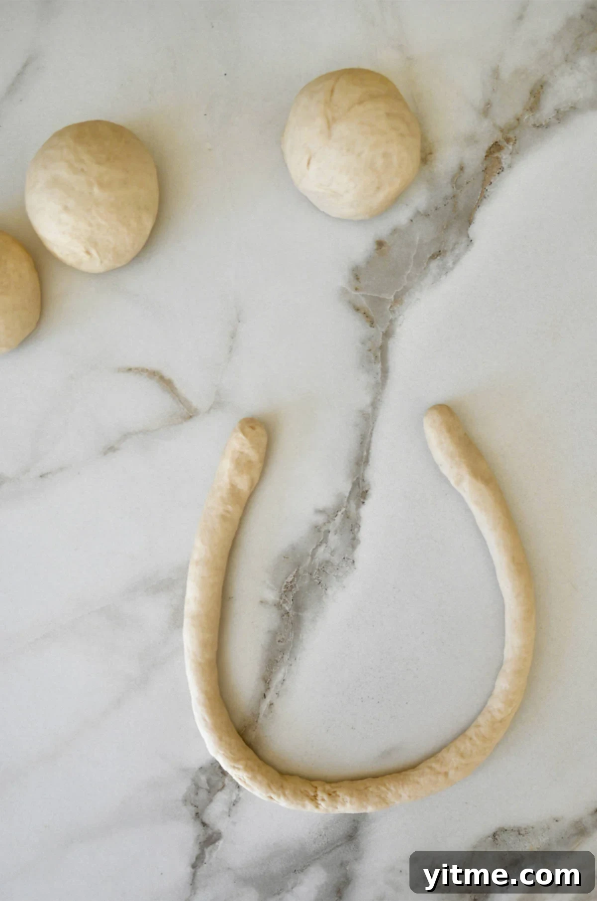 A long rope of dough in a "u" shape on a marble surface next to three balls of dough.
