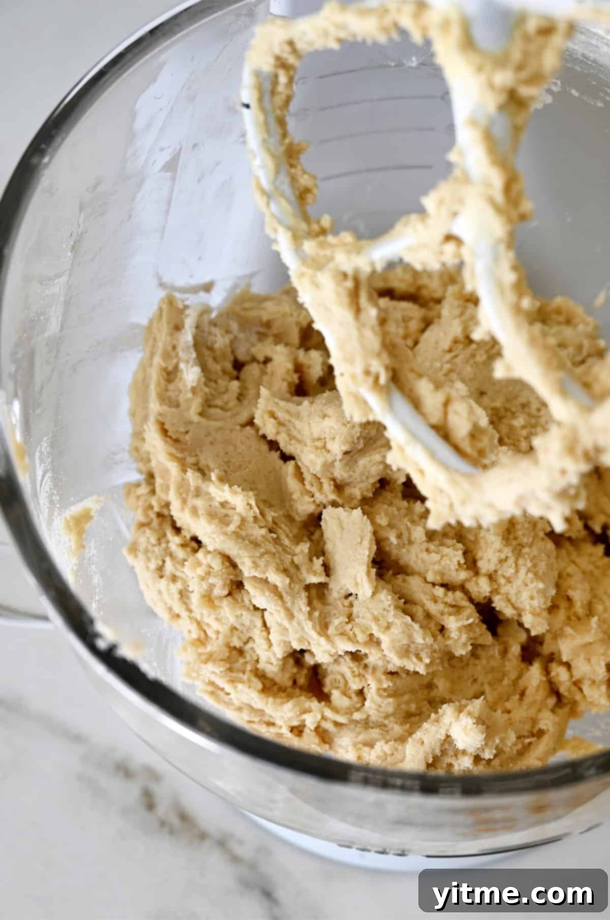 Sugar cookie dough covering the paddle attachment of a stand mixer above the glass bowl of the mixer containing more cookie dough.