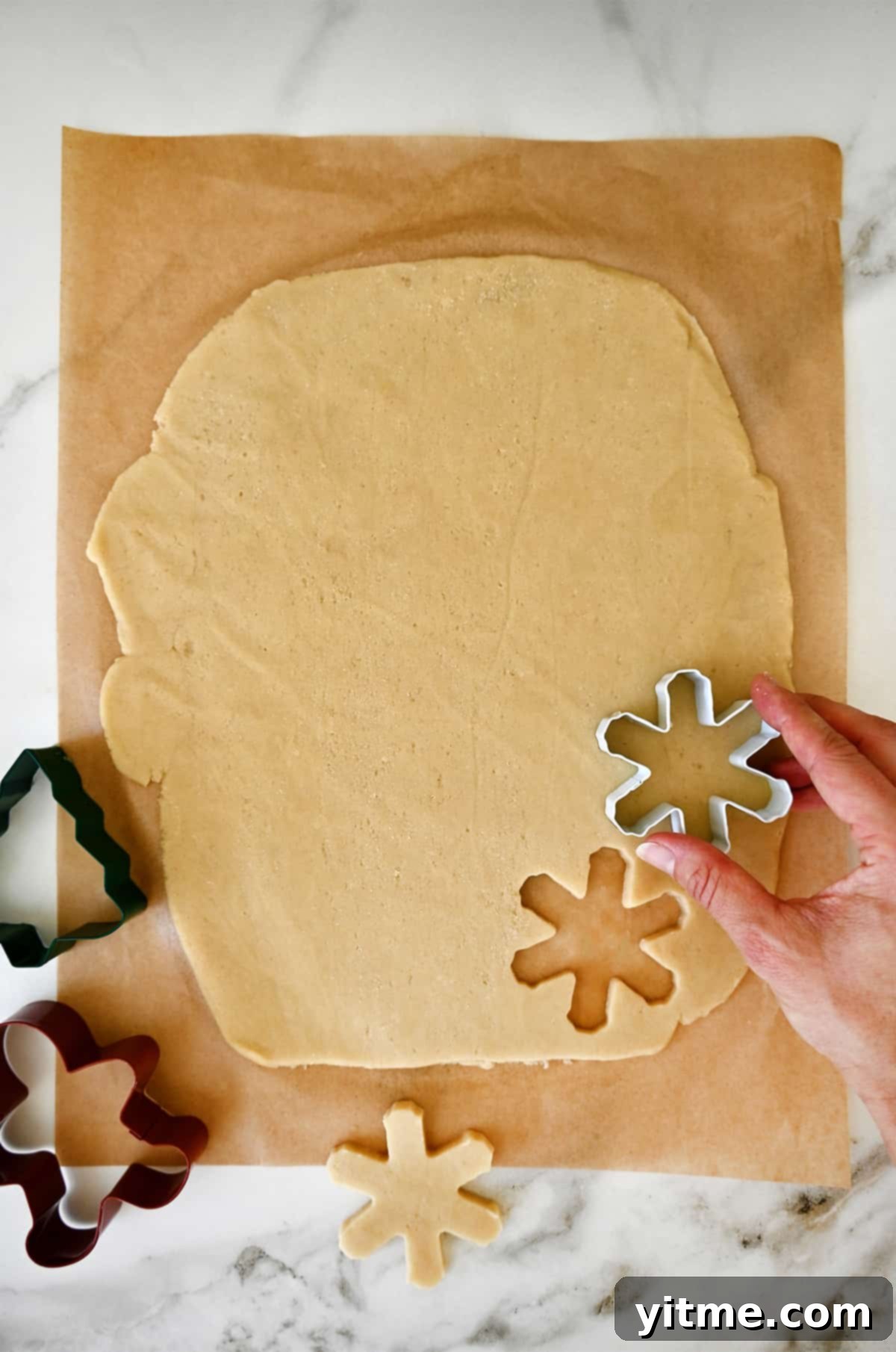 Rolled out dough on a sheet of parchment paper being cut into snowflake shapes with a gingerbread man and Christmas tree cookie cutter next to it.