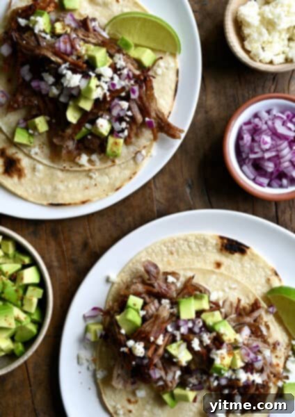 Balsamic Honey Pulled Pork: A Slow Cooker Delight 13 Two pulled pork tacos on separate plates. The tacos are topped with cubed avocado, crumbled cotija cheese and diced red onion. Small bowls with more toppings are beside the tacos.