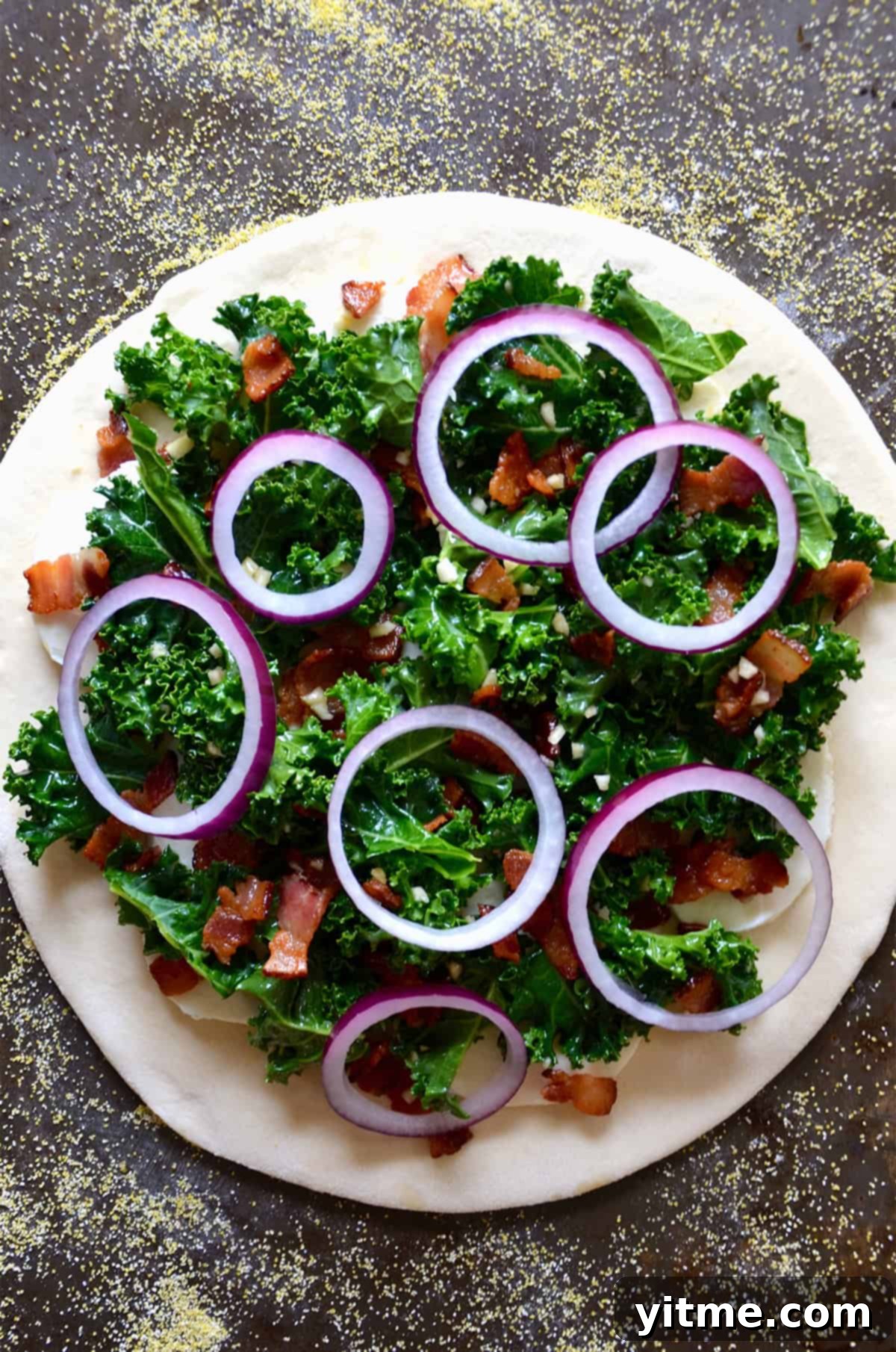 Garlic-Kissed Kale & Bacon Pie 6 A round of pizza dough topped with kale, chopped, crispy bacon and sliced red onion on a cornmeal-dusted surface.