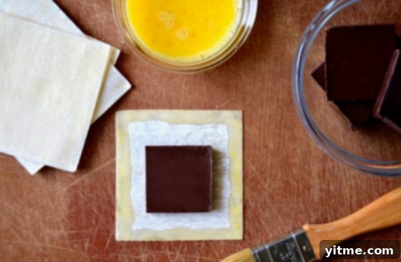 A well-organized cutting board featuring the essential ingredients for chocolate wontons: wonton wrappers, squares of chocolate, a bowl of egg wash, and a pastry brush.