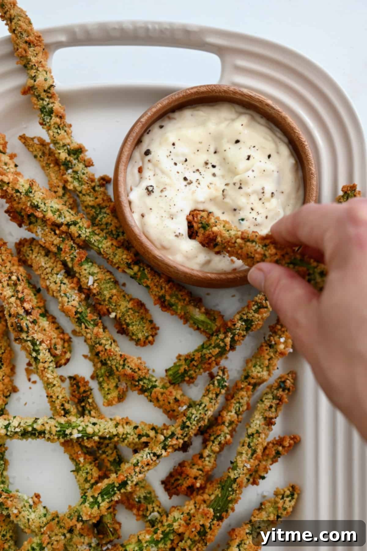 Baked asparagus fries piled on a white platter with a bowl of roasted garlic aioli. A hand dips a fry into the aioli.