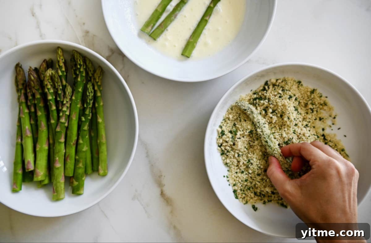 Three bowls holding asparagus, mayonnaise-egg white mixture, and breadcrumbs with parsley and parmesan for making asparagus fries.