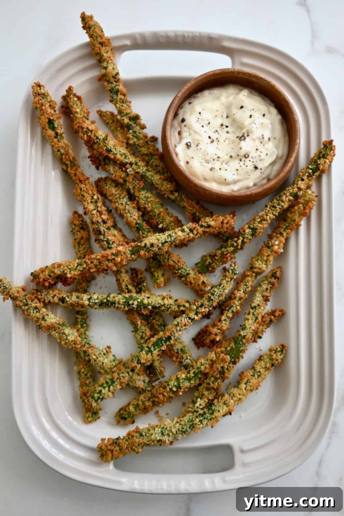 Baked asparagus fries piled on a white platter with a bowl of roasted garlic aioli.