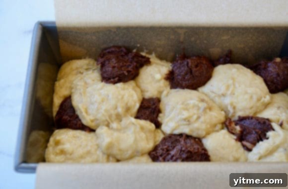 Bread pan lined with parchment paper, filled with alternating layers of banana batter and chocolate batter, ready for swirling