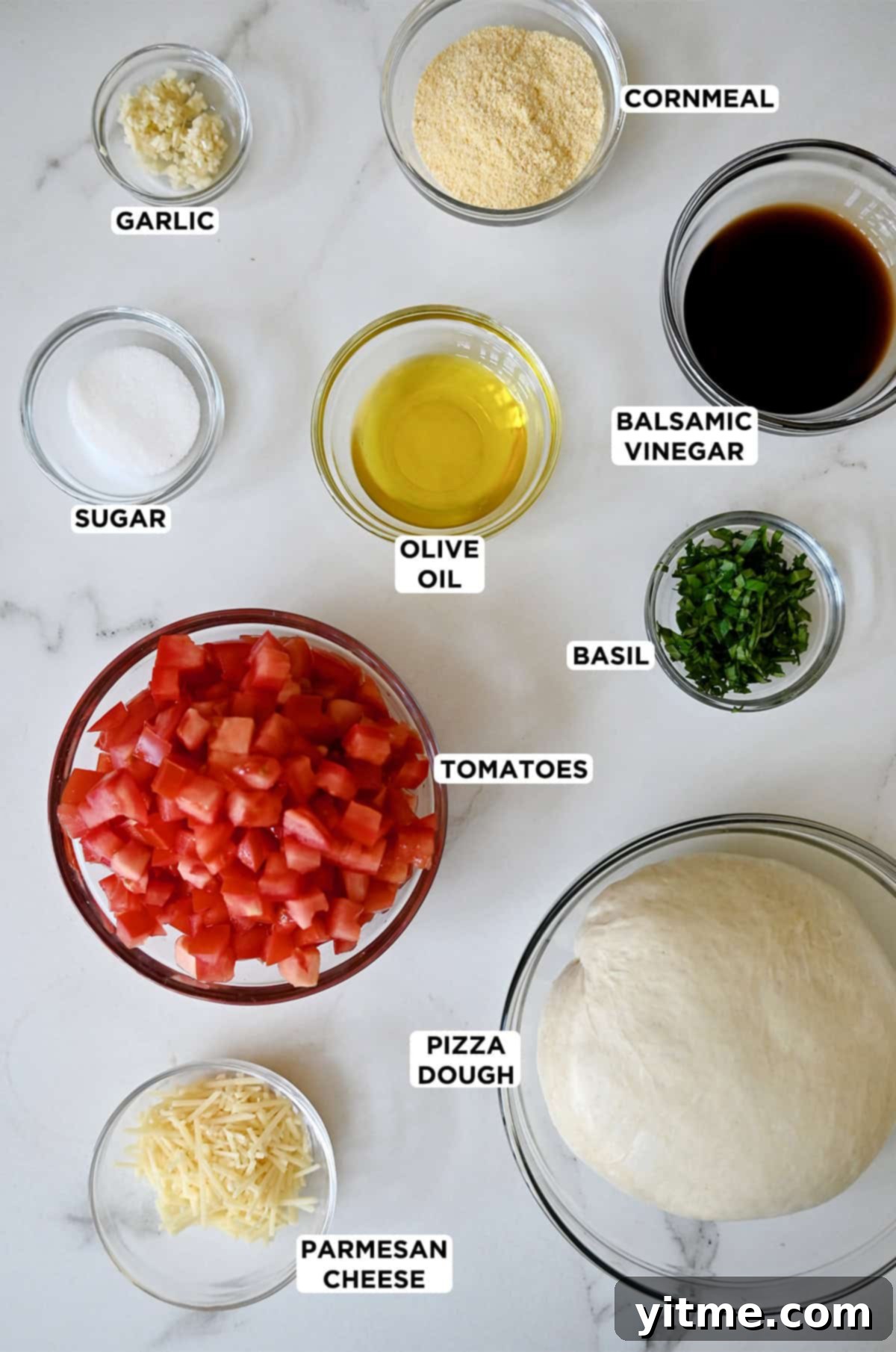 Various sizes of glass bowls containing pizza dough, minced garlic, diced tomatoes, olive oil, sugar, chopped fresh basil, balsamic vinegar, cornmeal and shredded Parmesan.