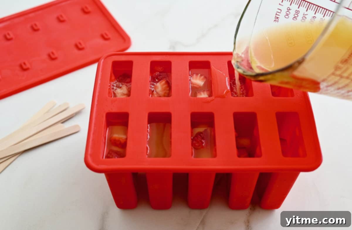 A hand holds a liquid measuring cup, precisely filling a multi-cavity popsicle mold with the vibrant peach sangria mixture.