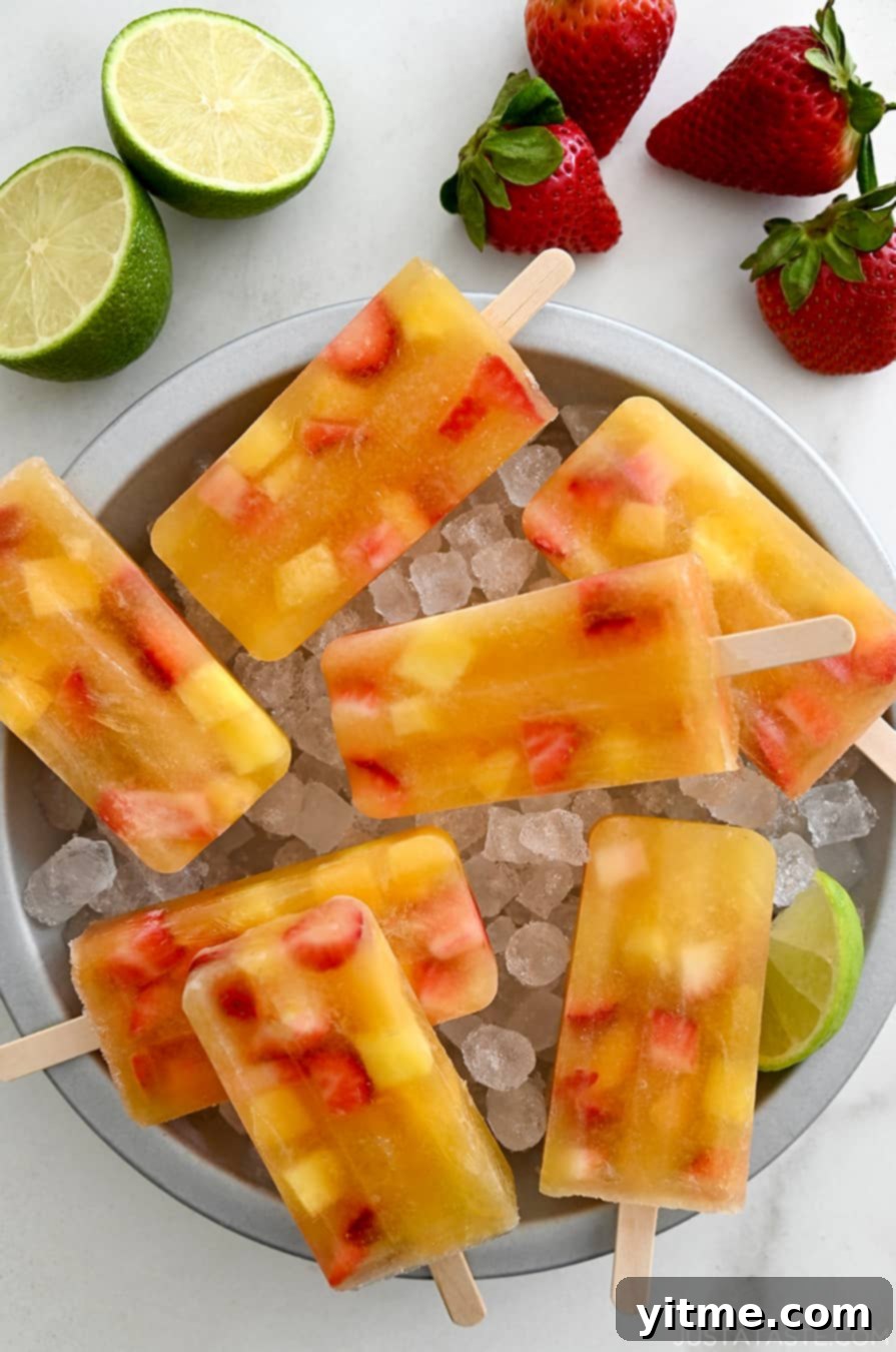 A top-down view of Peach Sangria Popsicles over ice in a pie plate next to fresh strawberries and a lime cut open in half, perfect for a summer dessert.