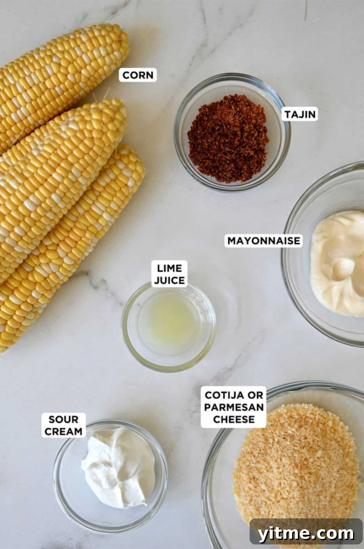 Essential ingredients laid out for making Mexican street corn: fresh sweet corn on the cob, creamy mayonnaise, tangy sour cream, crumbly Cotija cheese, zesty lime juice, and vibrant chili powder.
