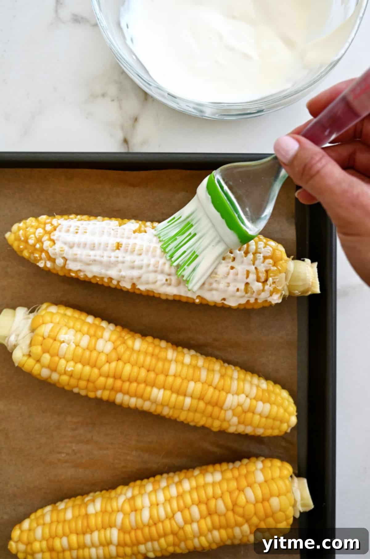 A hand using a silicone pastry brush to generously slather boiled corn cobs with a creamy and tangy elote sauce, preparing them for the next delicious step.