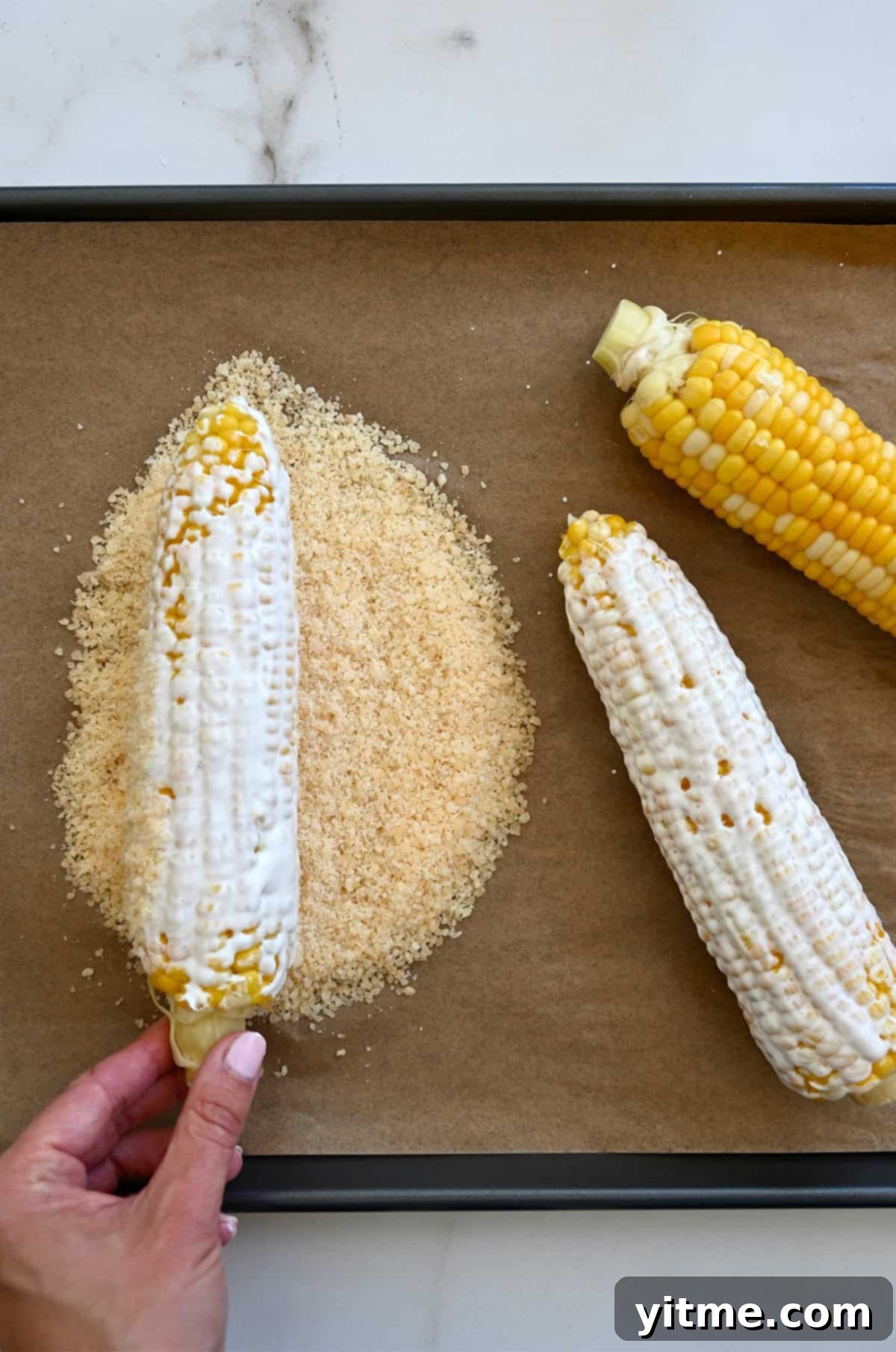 A close-up of a hand rolling a freshly cooked ear of corn, already coated in the creamy elote sauce, through a bowl of crumbled Cotija cheese for Mexican street corn.