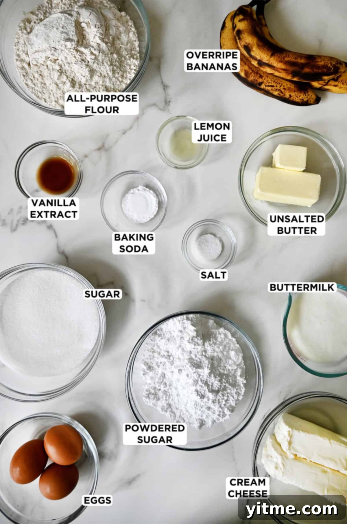 Banana cake and cream cheese frosting ingredients in various sizes of glass bowls: flour, overripe bananas, butter, lemon juice, baking soda, salt, buttermilk, vanilla extract, sugar, eggs, powdered sugar and cream cheese.