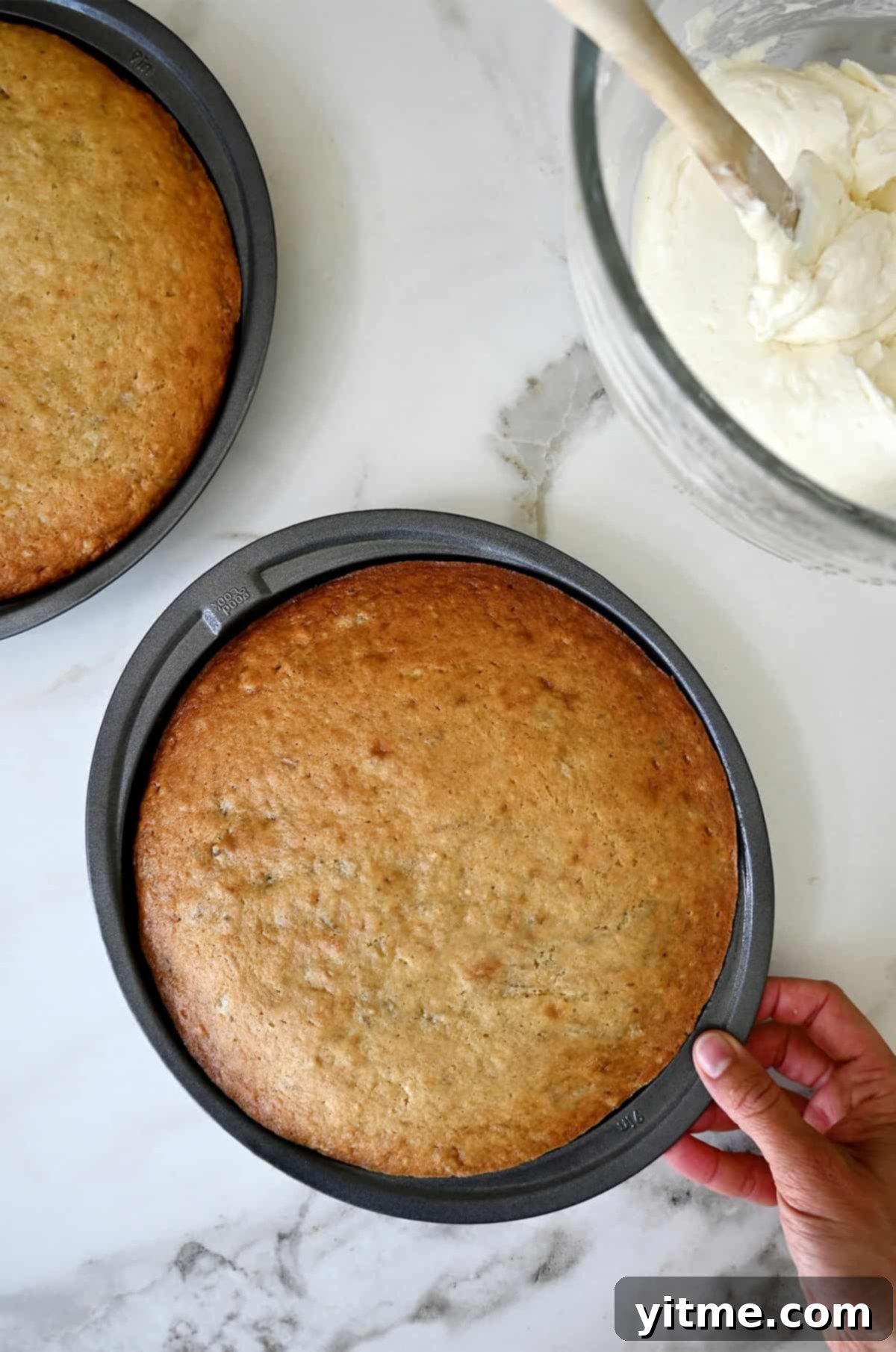 Baked moist banana cake in two round cake pans next to a bowl containing cream cheese frosting.