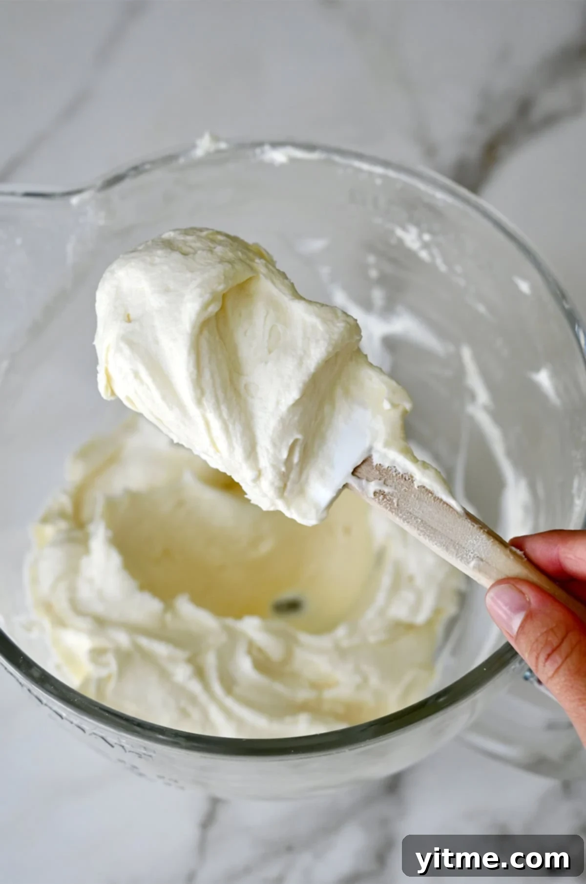 A hand holds a spatula covered in whipped cream cheese frosting.