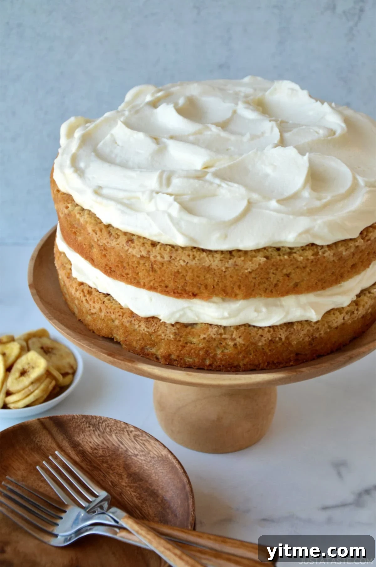 A two-layer banana cake with a thick layer of cream cheese frosting in the middle and on the top, garnished with chopped walnuts, on a wooden cake stand.