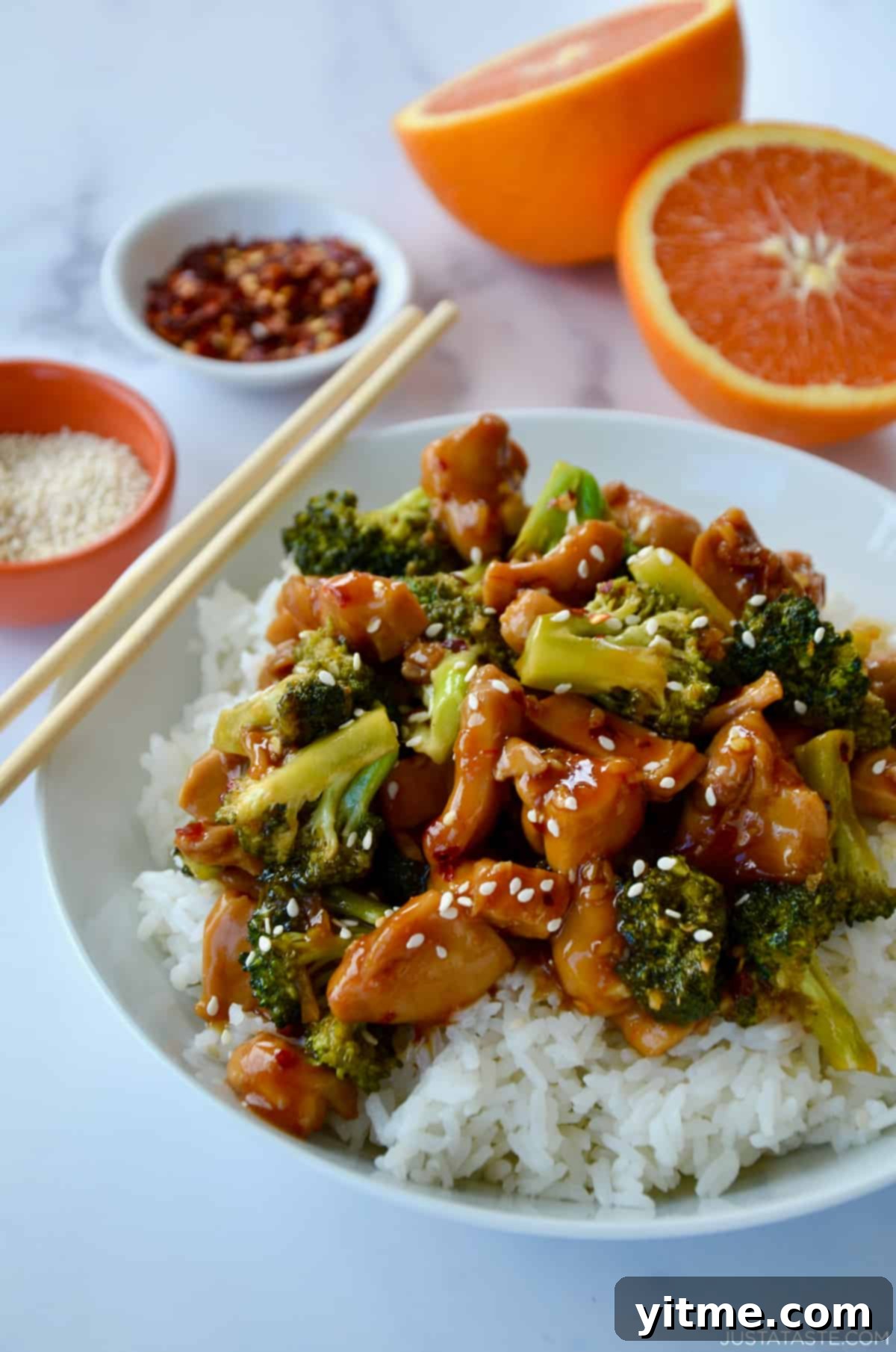 Orange chicken and broccoli atop steamed white rice in a white bowl with chopsticks.