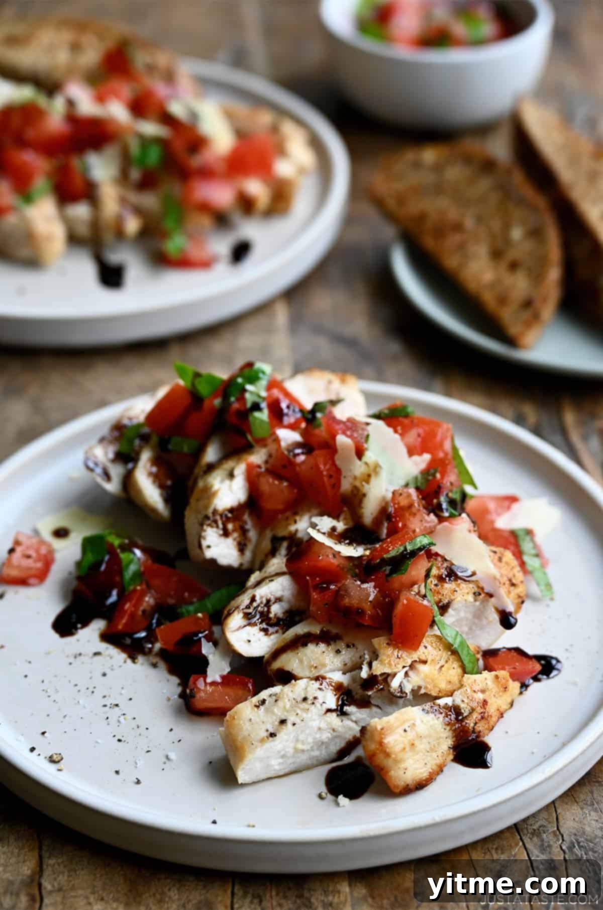 Easy bruschetta chicken topped with shaved Parmesan cheese and a drizzle of balsamic syrup on a white dinner plate. A plate piled high with crusty bread slices is nearby.