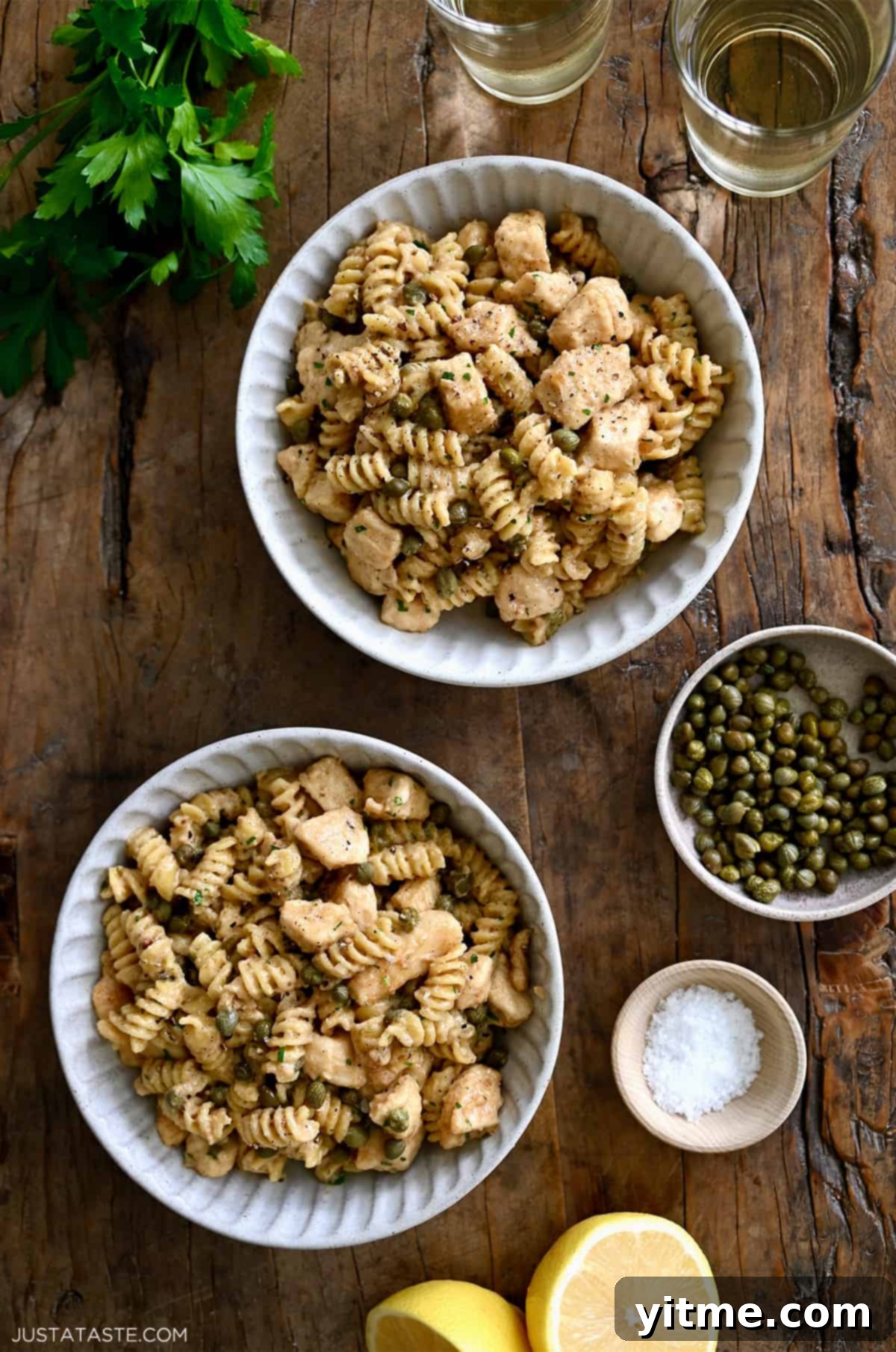 A top-down view of two bowls containing Chicken Piccata Pasta next to two small bowls containing capers and sea salt.