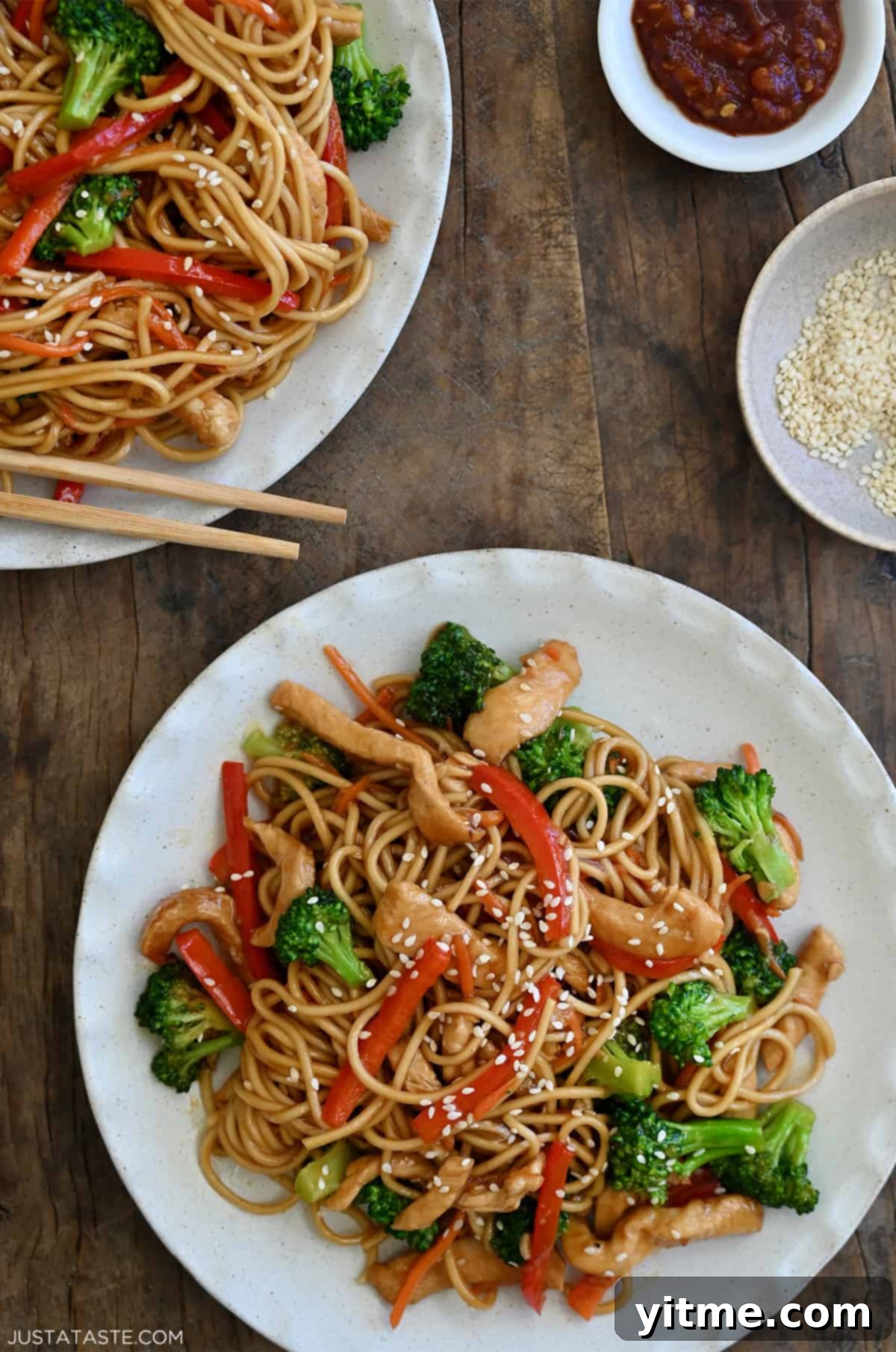 Two dinner plates containing Teriyaki Chicken Stir-Fry over noodles next to a small bowl filled with sesame seeds and a small bowl containing sambal oelek.
