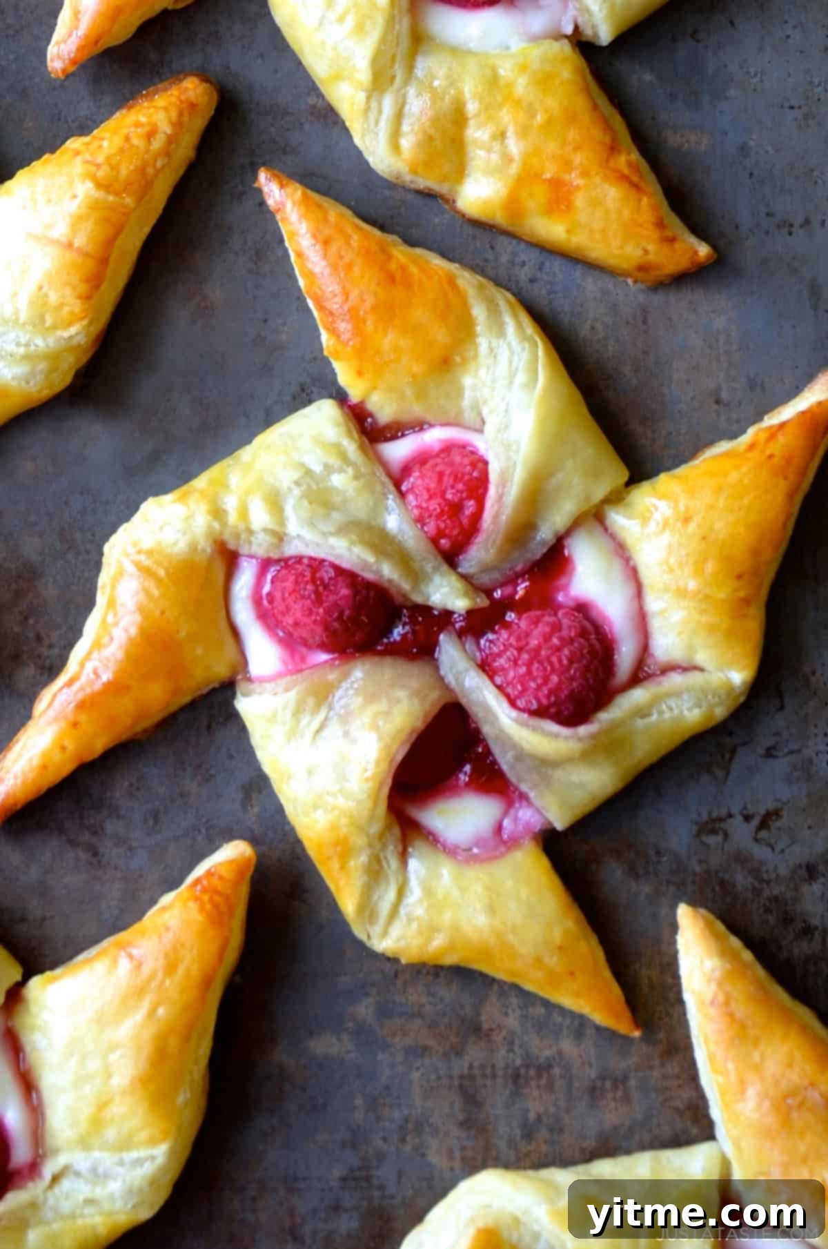 A single, perfectly golden brown Raspberry Cream Cheese Pinwheel Pastry, with its flaky layers and vibrant fruit filling visible, on a dark background.