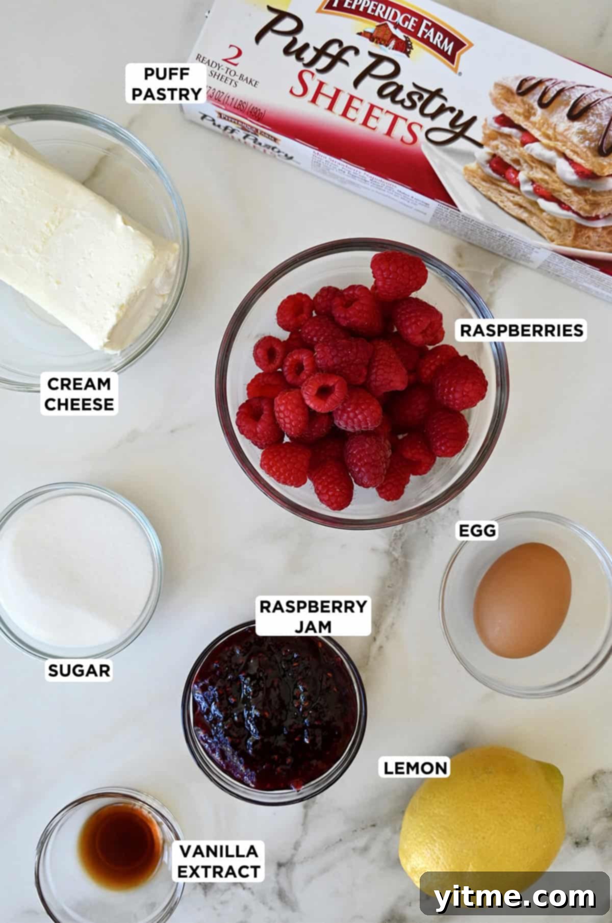 A collection of ingredients for raspberry cream cheese pastries, including frozen puff pastry sheets, a block of cream cheese, fresh raspberries, sugar, lemon, and vanilla extract.