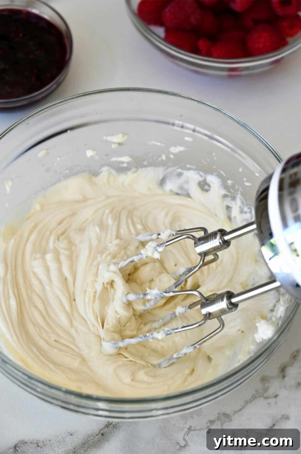 Cream cheese, sugar, lemon juice, lemon zest, and vanilla extract being blended in a bowl with a handheld electric mixer until smooth and creamy.