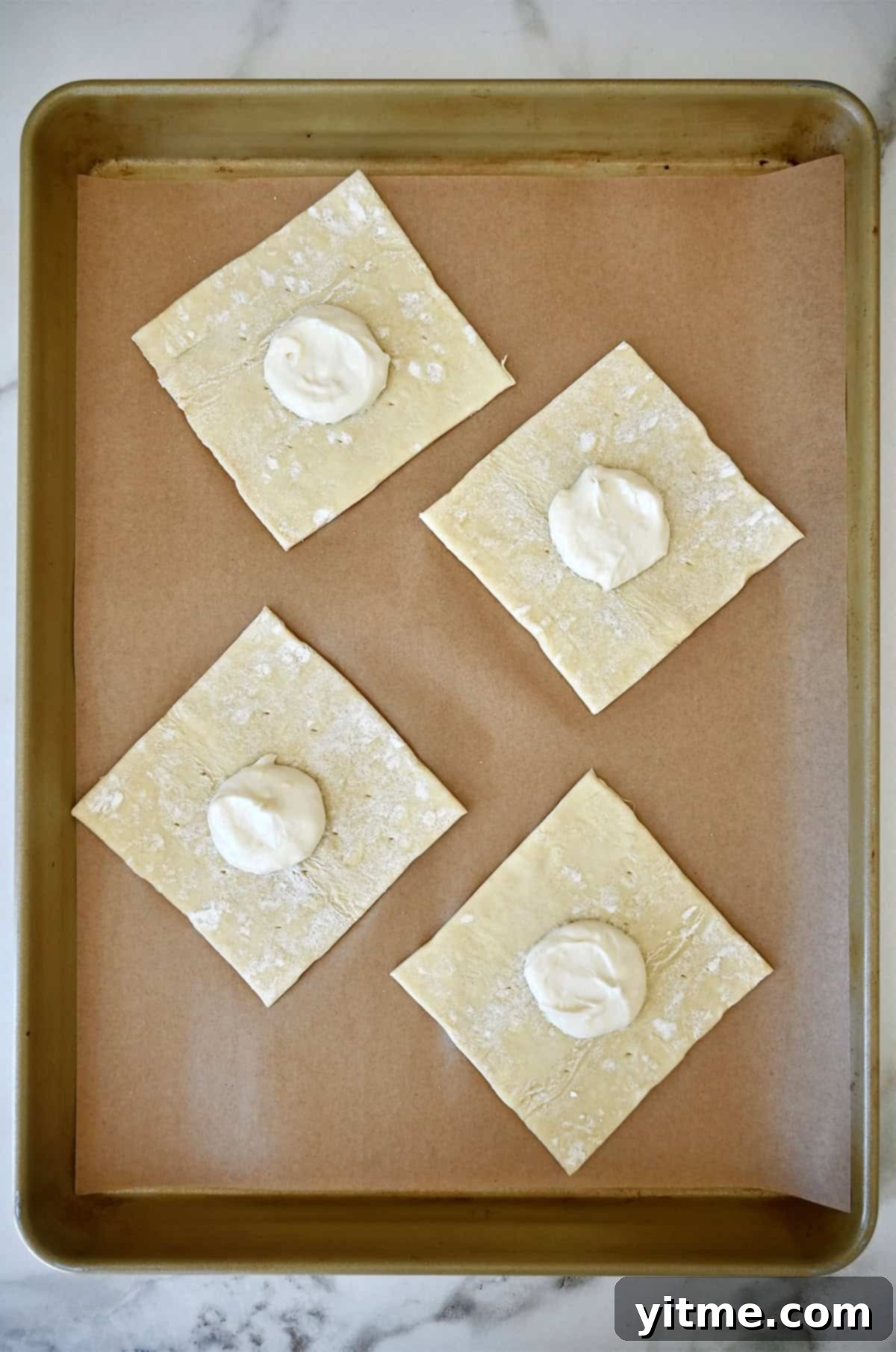 Four squares of puff pastry on a parchment-lined baking sheet, each topped with a dollop of creamy lemon cream cheese filling.