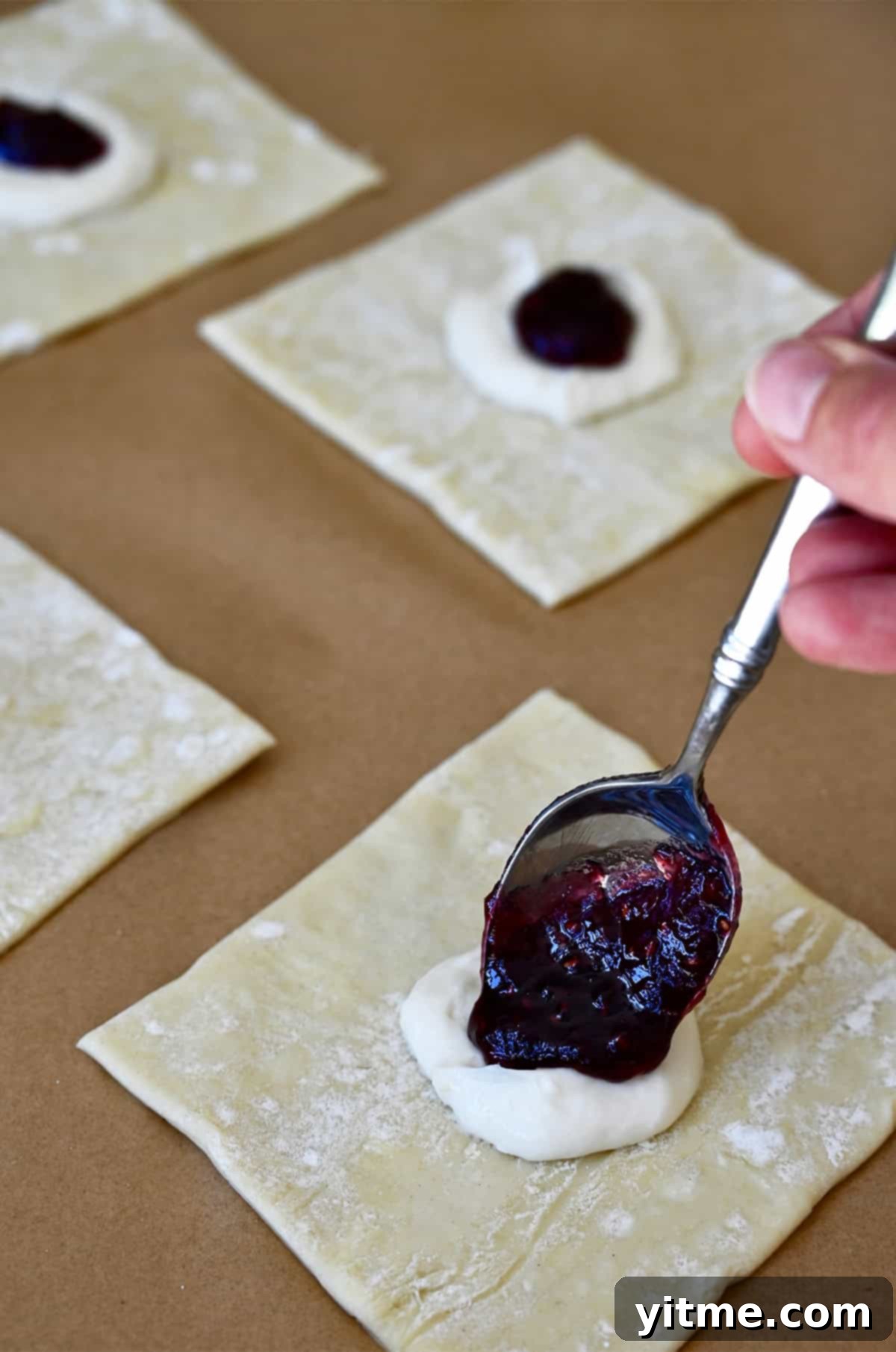 A spoon adding a dollop of vibrant raspberry jam on top of the cream cheese filling on a puff pastry square.