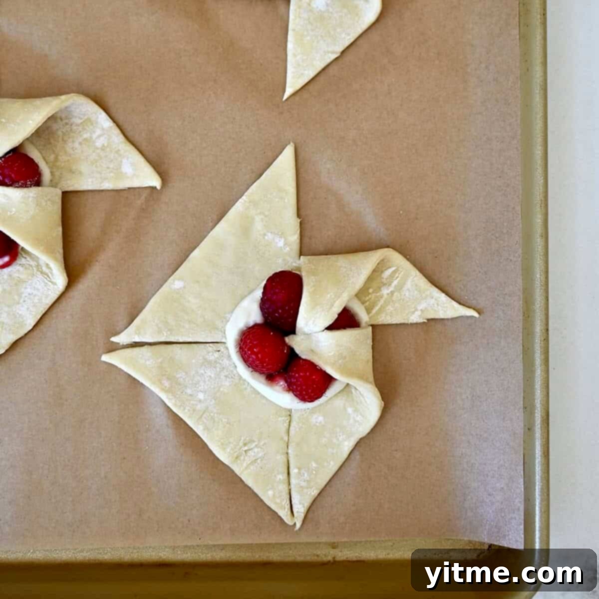 Step 2 of shaping a pinwheel: Folding a second corner of the puff pastry over the filling, beginning to form the pinwheel.