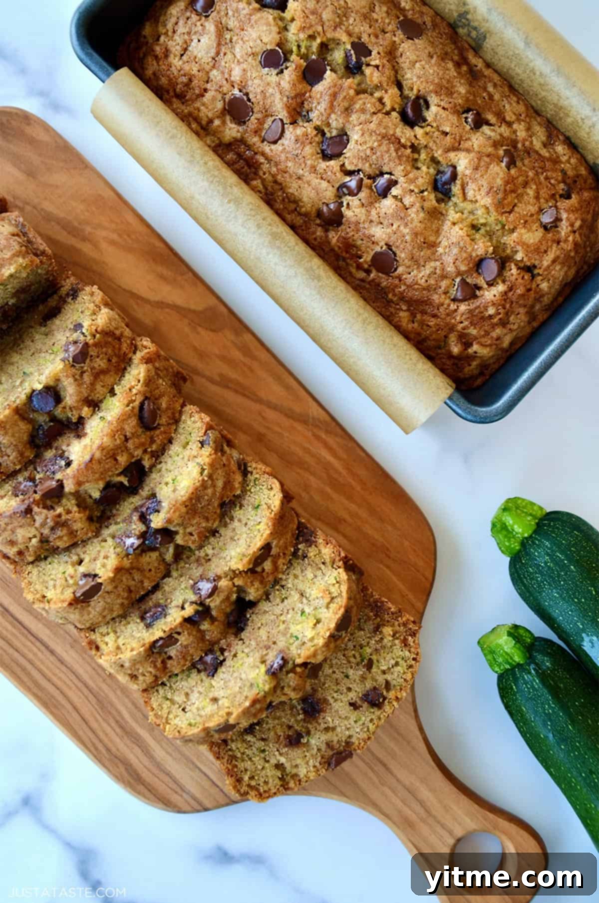 Two loaves of moist chocolate chip zucchini bread, one in a loaf pan and one cut into perfect slices on a cutting board.