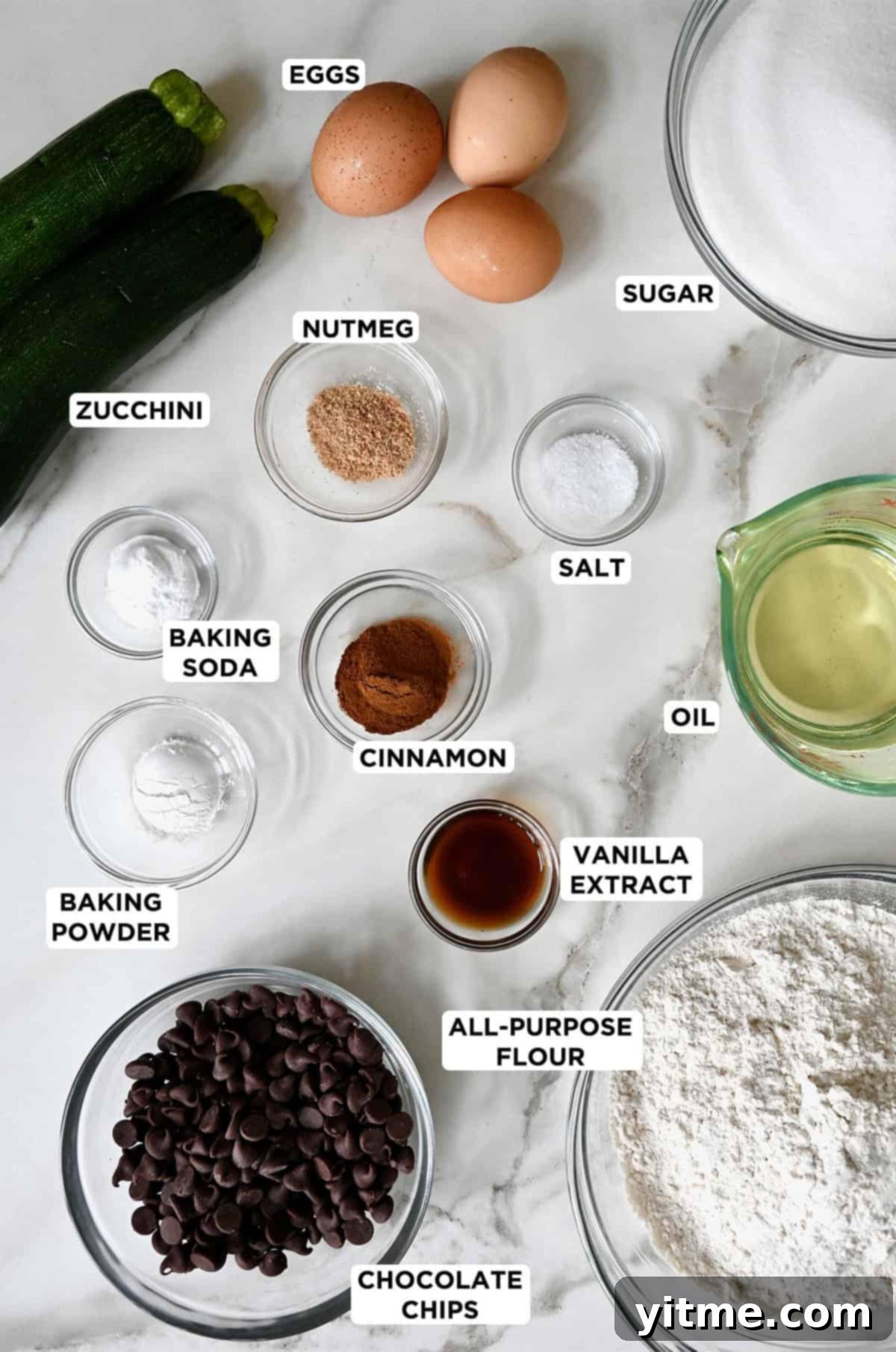 Ingredients for zucchini bread with chocolate chips in glass bowls, including eggs, sugar, nutmeg, cinnamon, salt, baking soda, vanilla extract, baking powder, flour, chocolate chips and two zucchini.