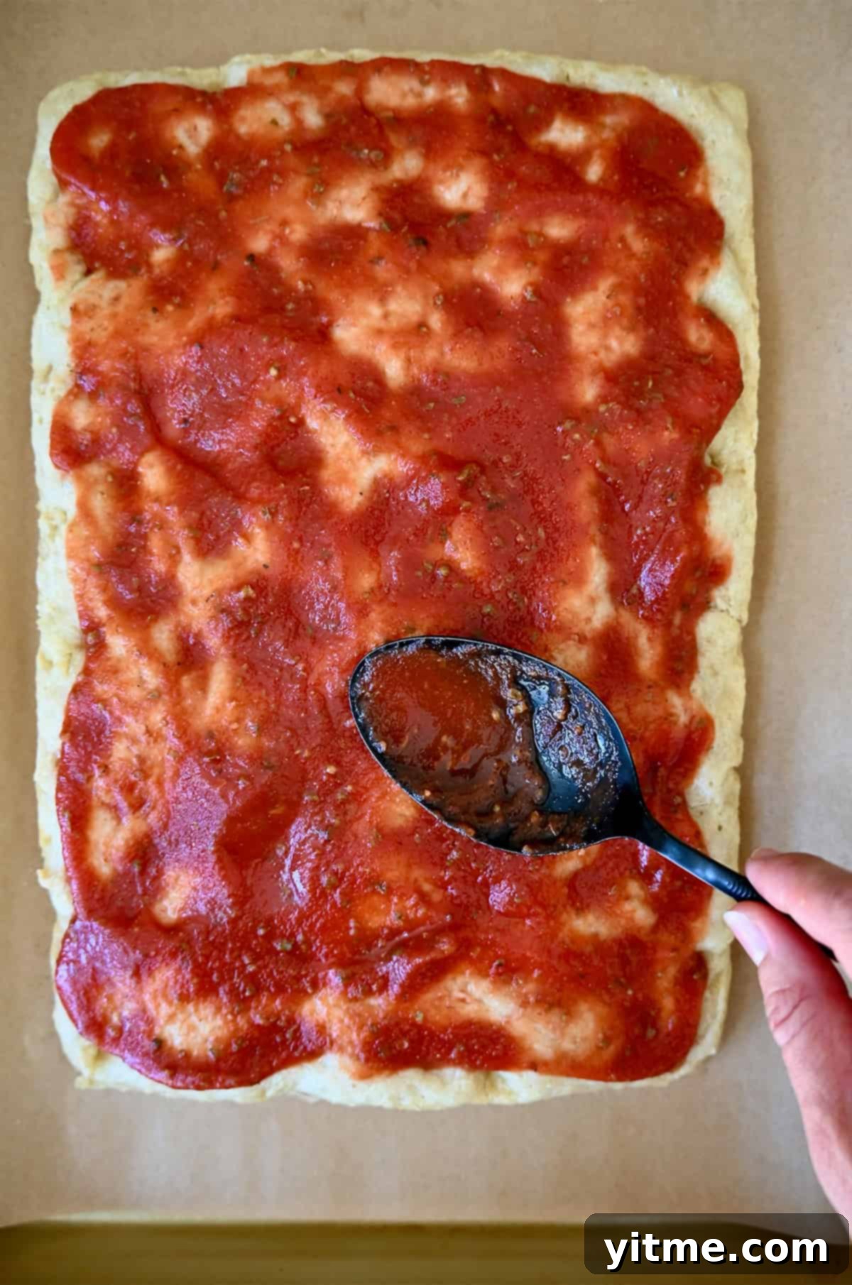 The back of a spoon spreads marinara sauce atop focaccia bread.