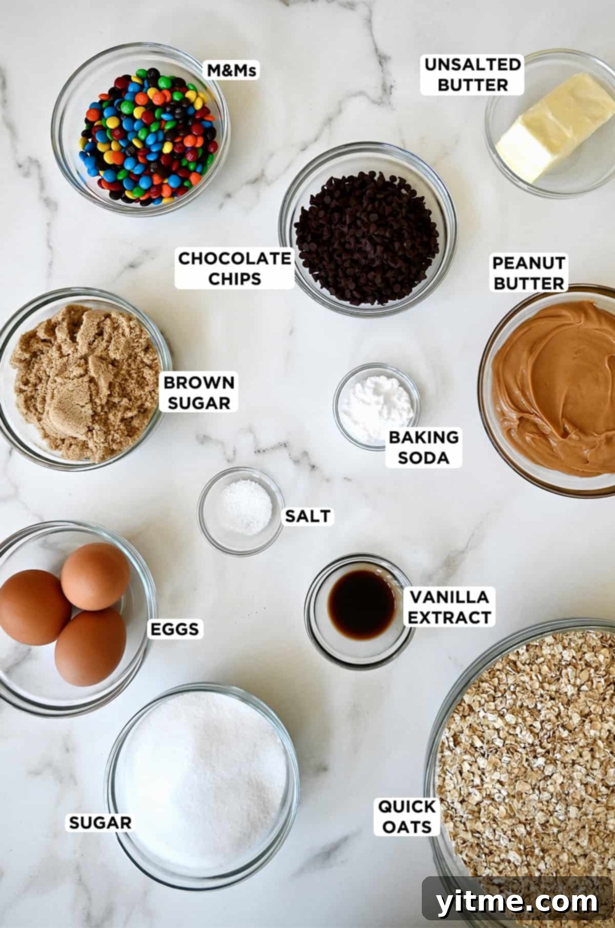 Glass bowls containing ingredients to make monster cookie bars, including M&Ms, chocolate chips, baking soda, white and brown sugars, vanilla extract, peanut butter, eggs and old-fashioned oats, all neatly arranged.