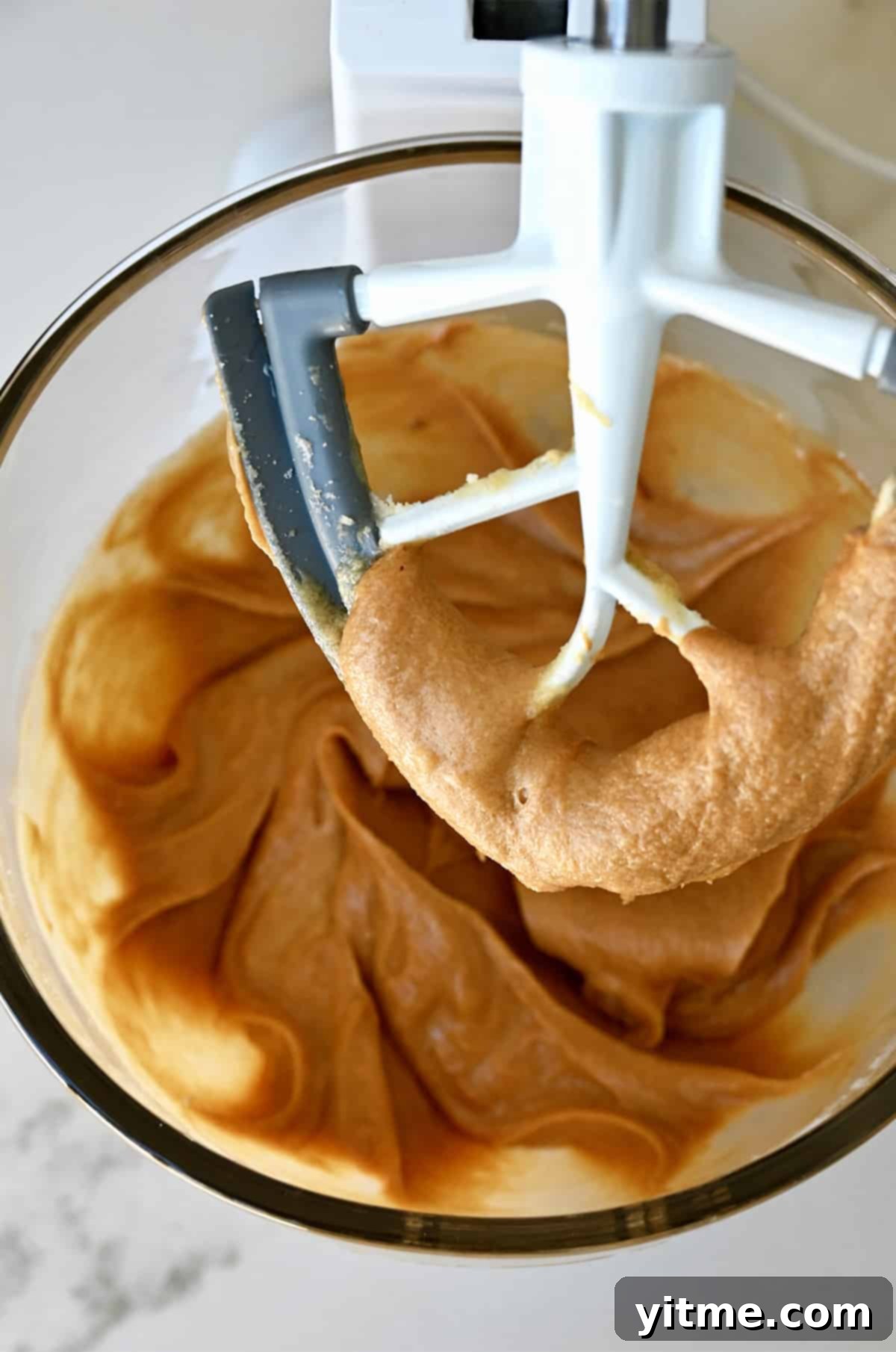 The paddle attachment of a stand mixer coated in a sugar and peanut butter mixture above a bowl containing the same mixture, showing the smooth texture achieved.