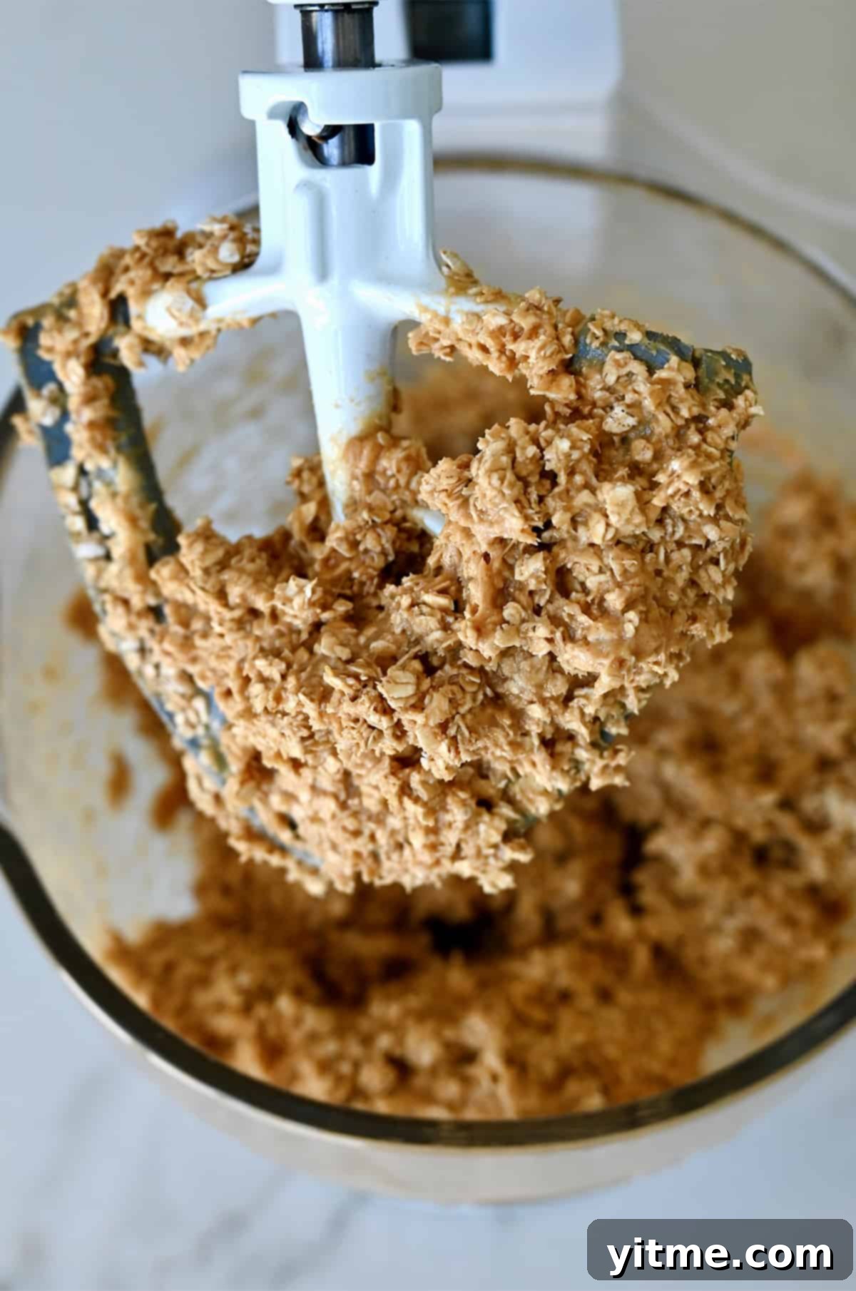 An oatmeal peanut butter mixture covering the paddle attachment of a stand mixture, demonstrating the thick, rich consistency of the dough.