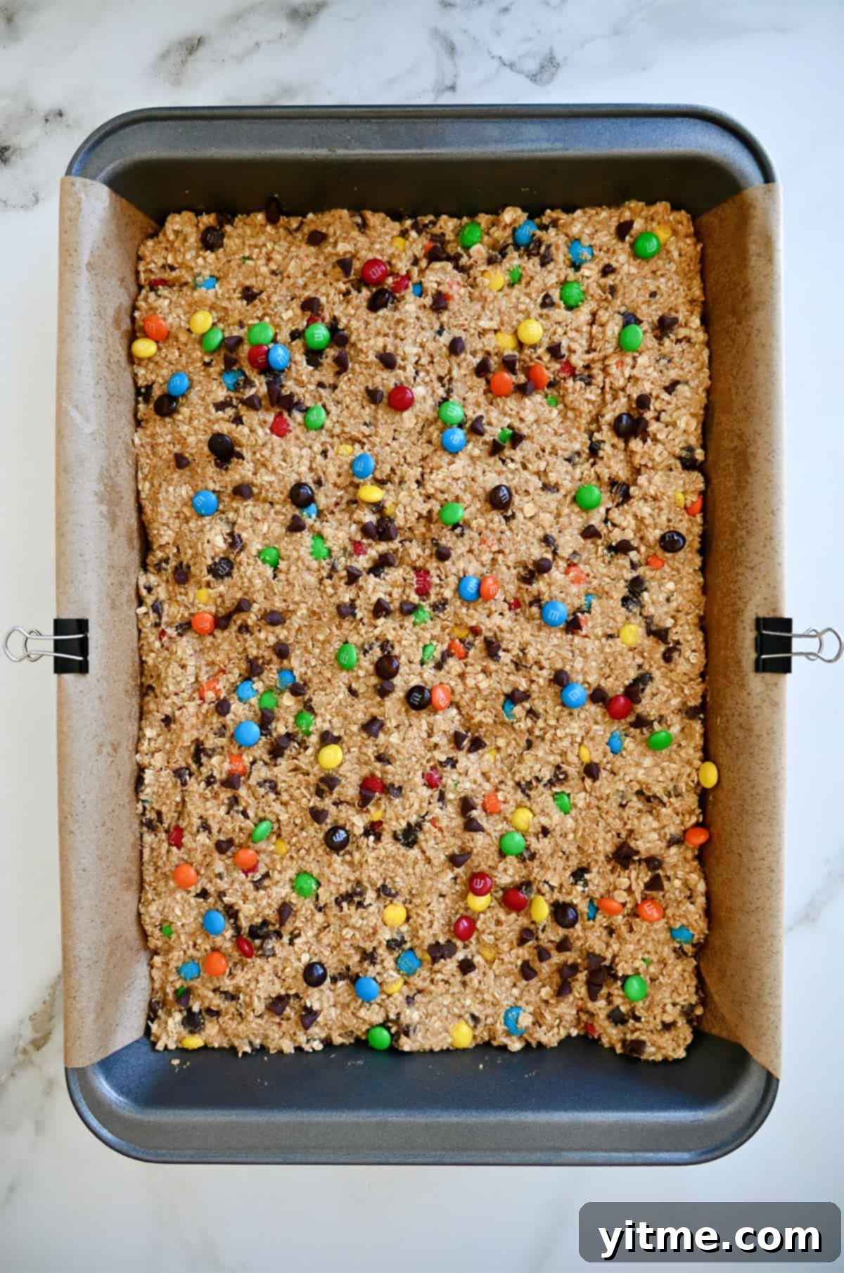 Monster cookie dough spread into an even layer in a 9x13 baking pan lined with parchment paper and secured with metal binder pins, ready for baking.