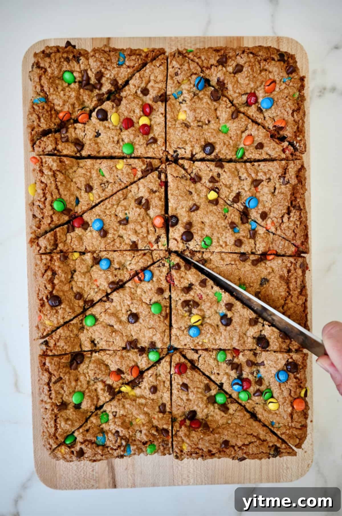 A hand holding a sharp knife cuts peanut butter oatmeal bars studded with M&Ms and chocolate chips into perfect triangles, highlighting the easy slicing.