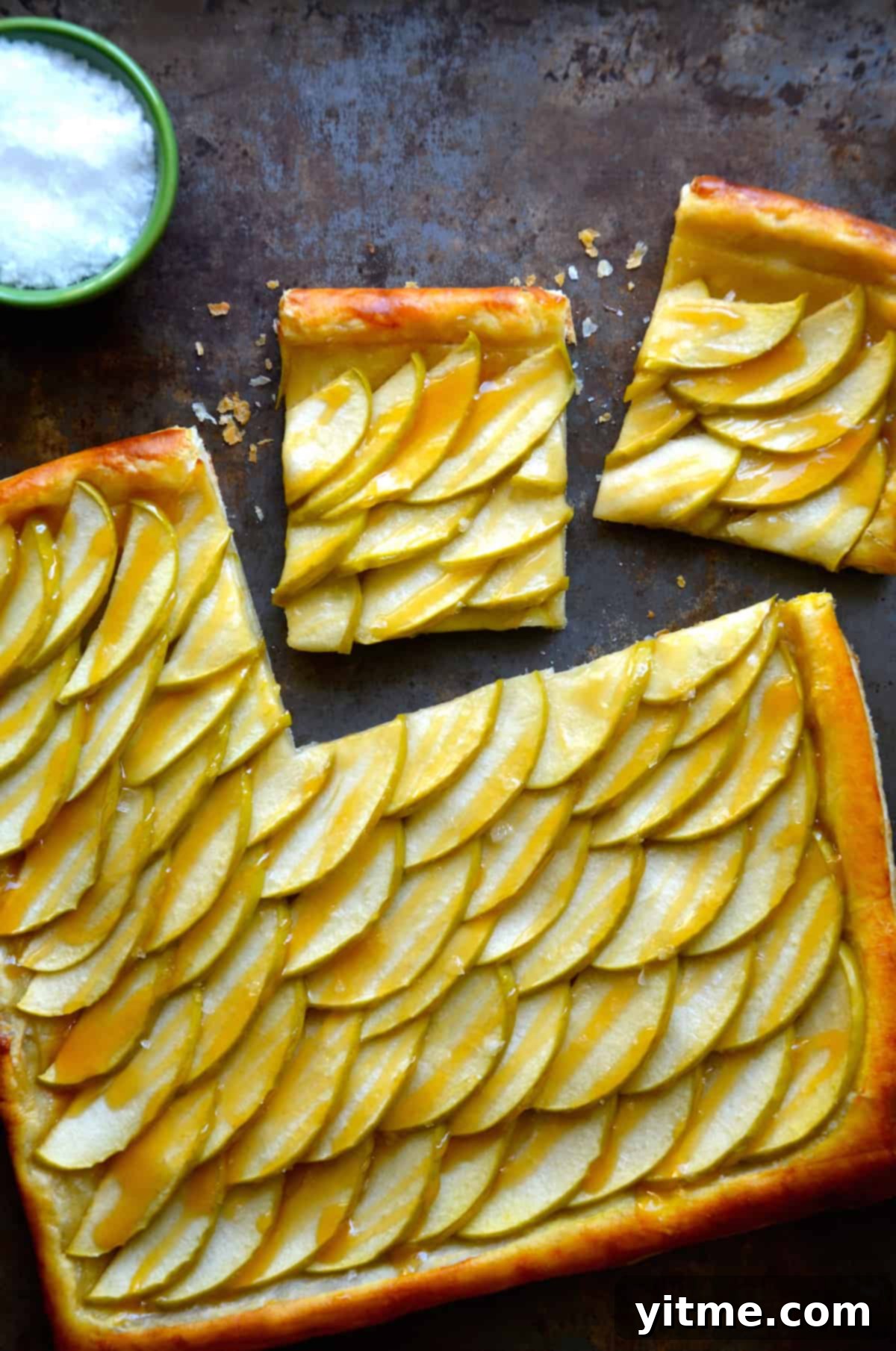 A close-up of a salted caramel apple tart made with puff pastry, featuring sliced portions and a salt bowl. 