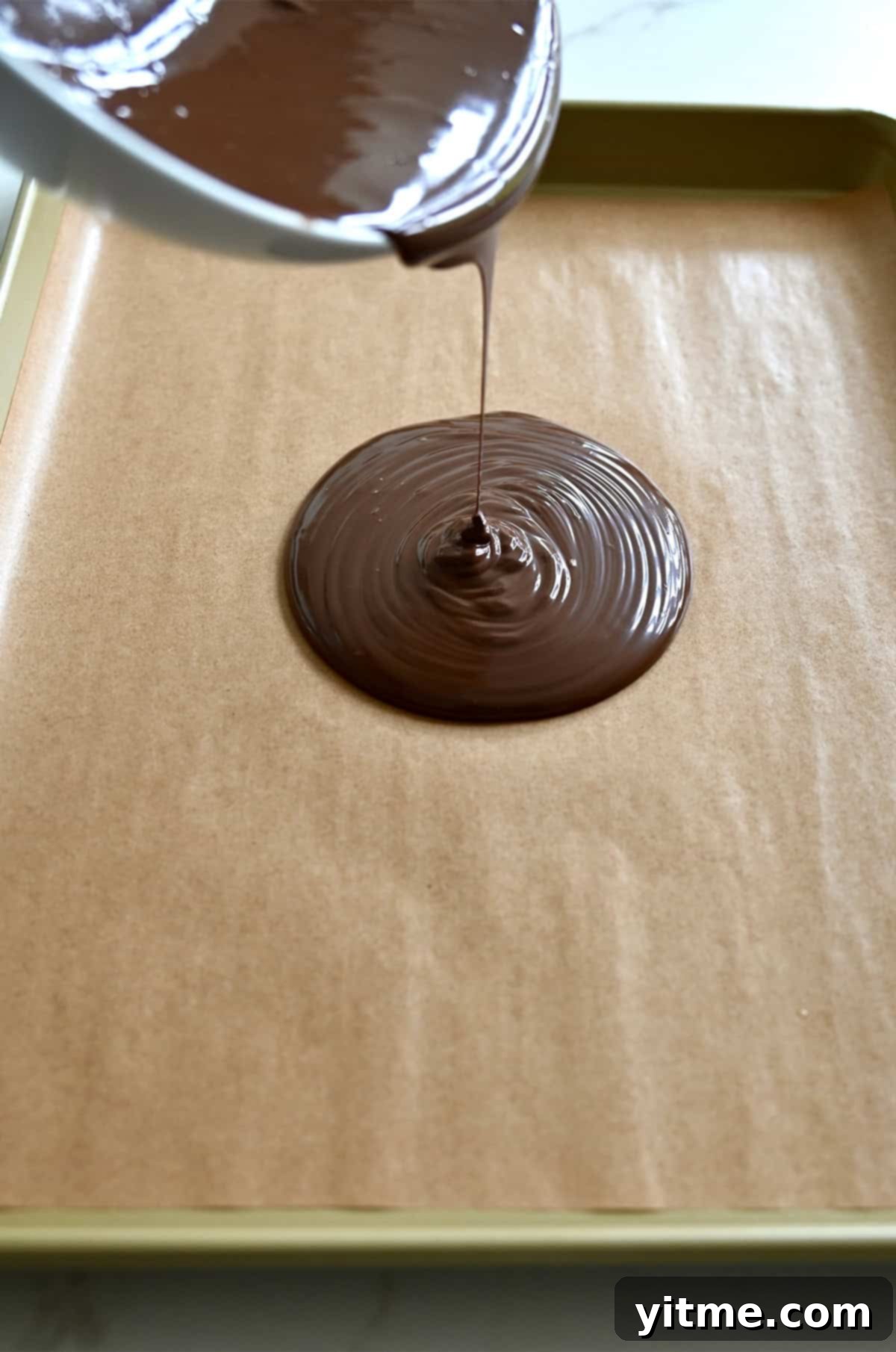 Melted chocolate is being poured from a bowl onto a parchment paper-lined baking sheet.