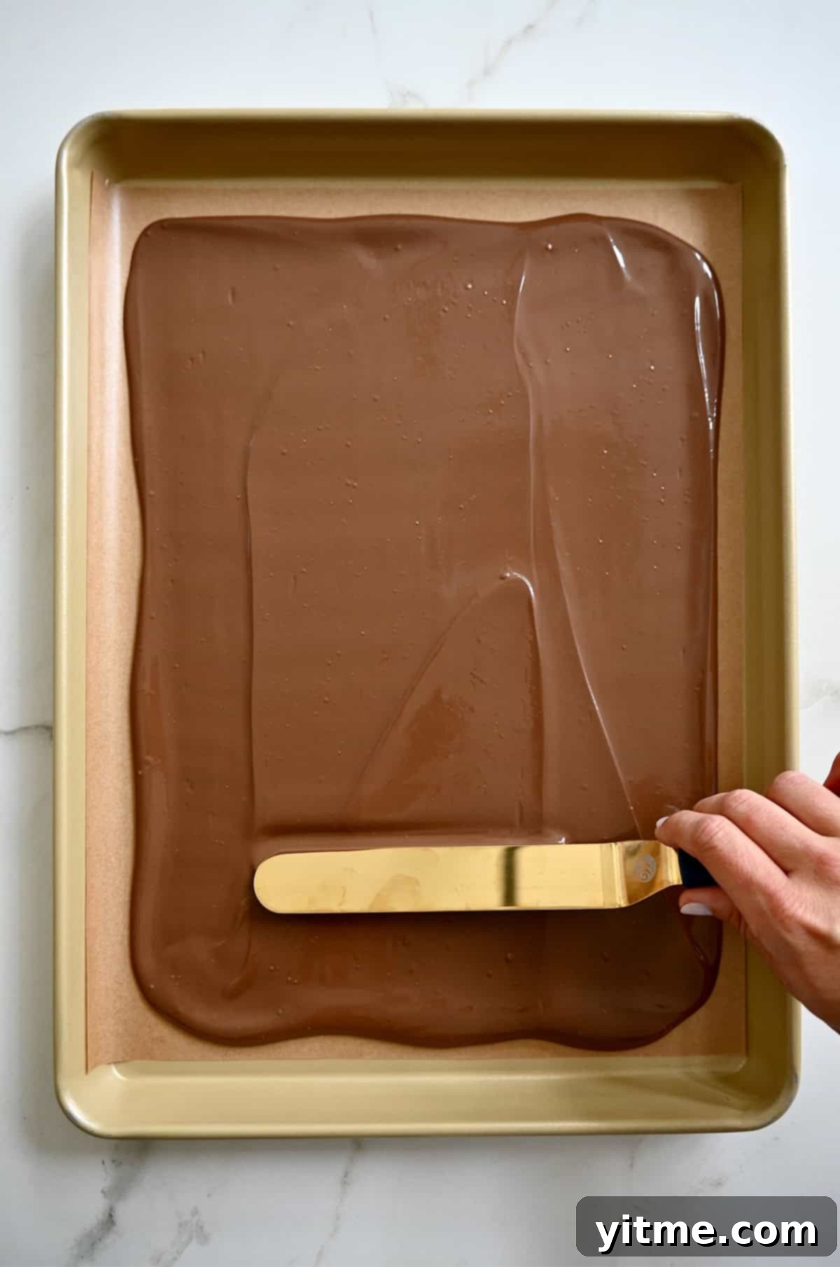 A hand holding an offset spatula spreads melted chocolate atop a parchment paper-lined baking sheet to make chocolate bark.