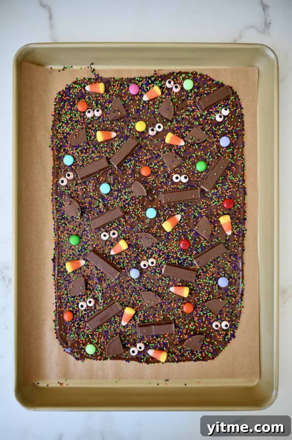 Chocolate bark topped with sprinkles, chopped candy bars, candy corn, M&Ms and bloodshot candy eyes is cooling on a parchment paper-lined baking tray.