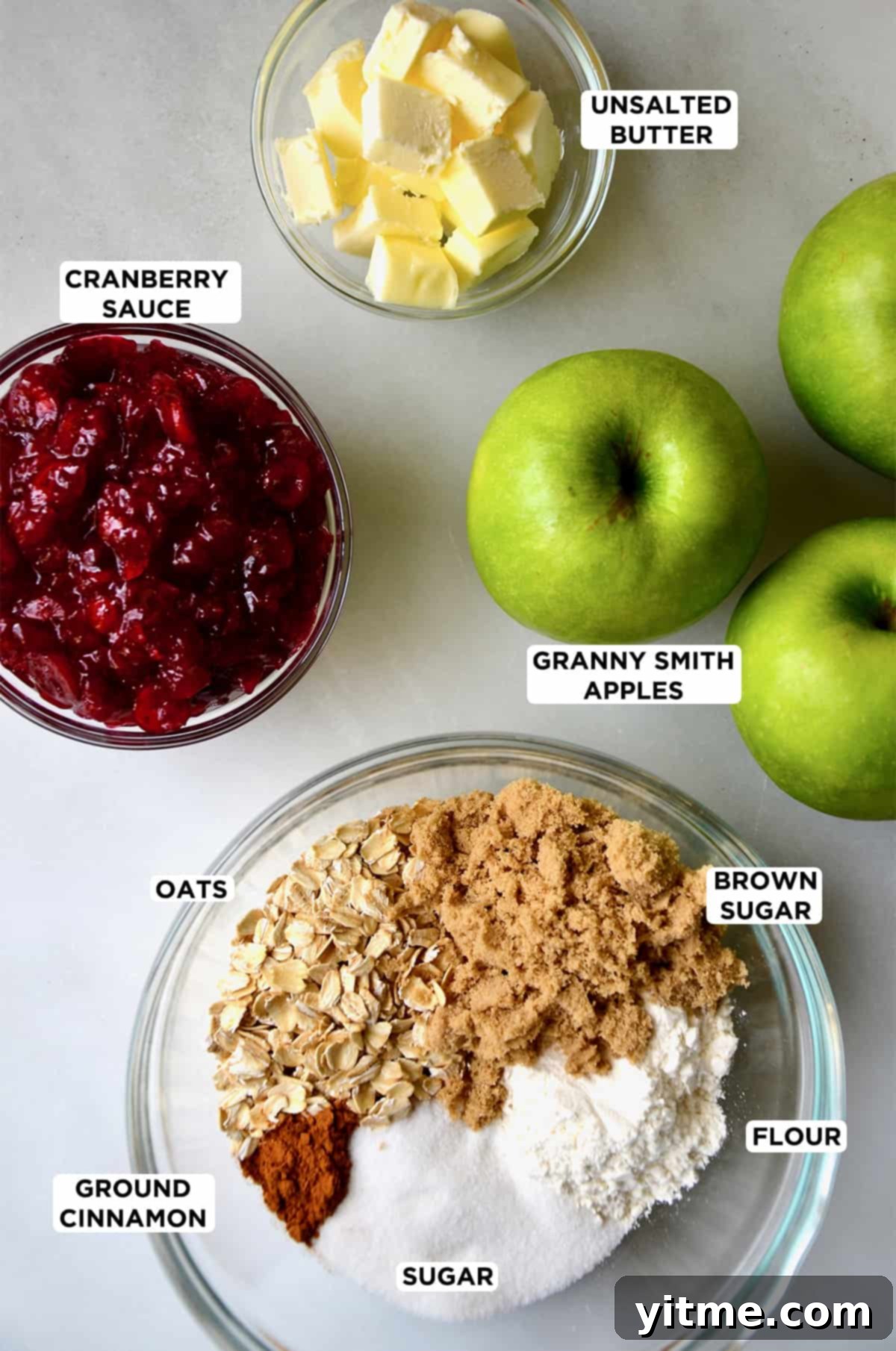 Granny Smith apples next to a bowl containing cranberry sauce, a bowl with cubed butter, and a bowl containing oats, brown sugar, flour and spices.