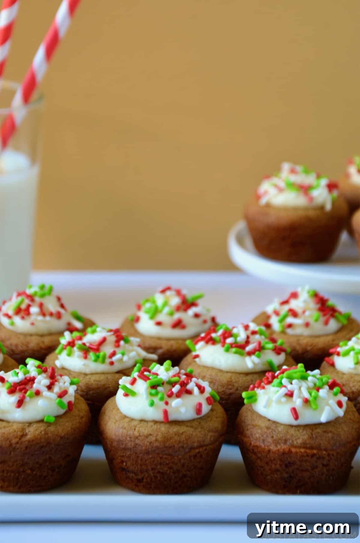 Gingerbread cookie cups topped with creamy cream cheese frosting and colorful sprinkles, displayed on a white platter with a glass of milk and more cookie cups in the background.