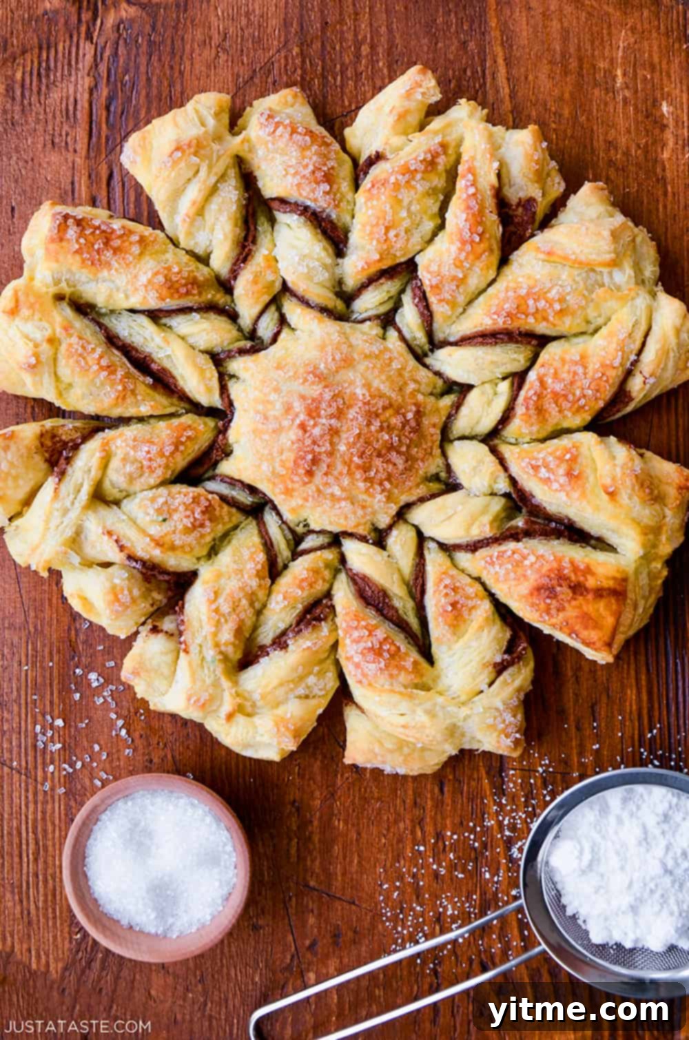 A top-down view of a Nutella Puff Pastry Snowflake, showcased next to a sifter filled with powdered sugar and a small bowl of sanding sugar.