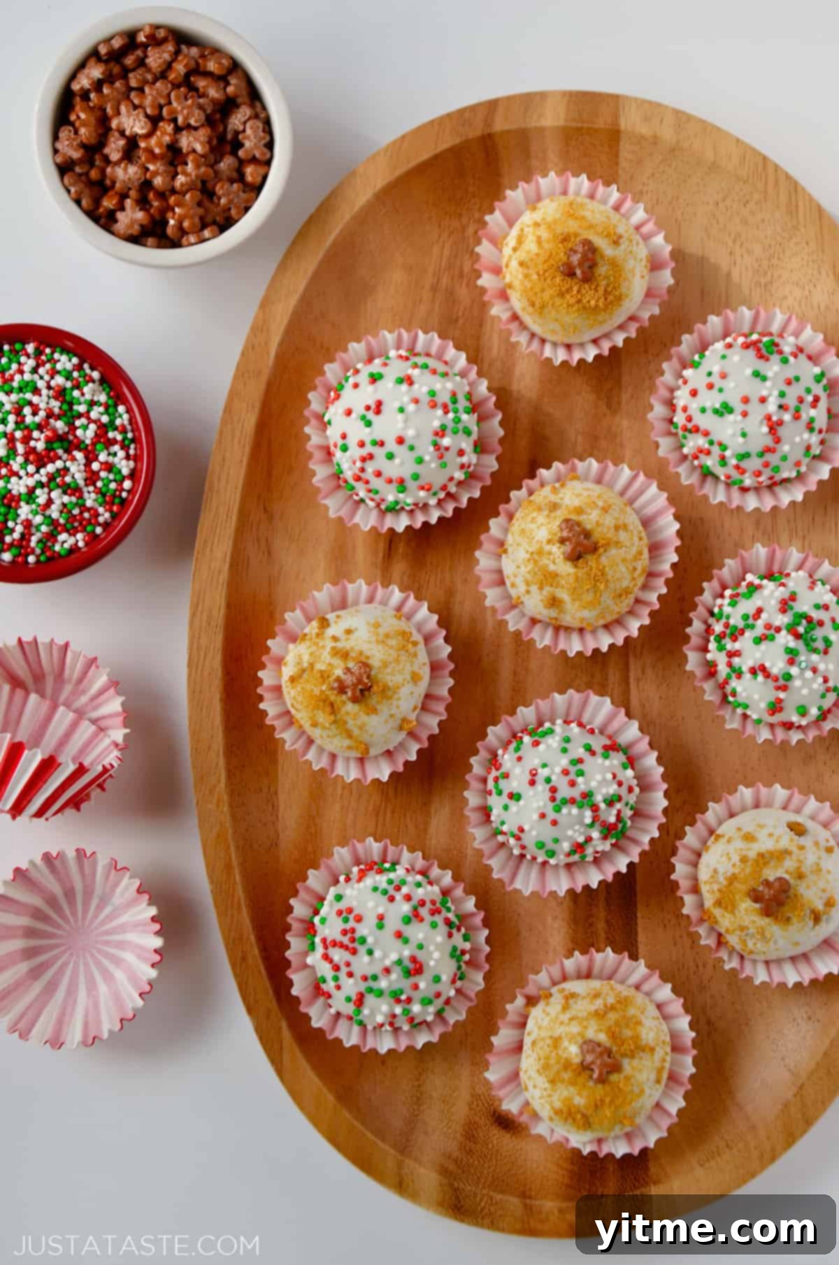 A wooden platter adorned with ten chocolate- and sprinkle-covered gingerbread cookie balls, elegantly wrapped in red-and-white striped wrappers, alongside more wrappers and small bowls of sprinkles.